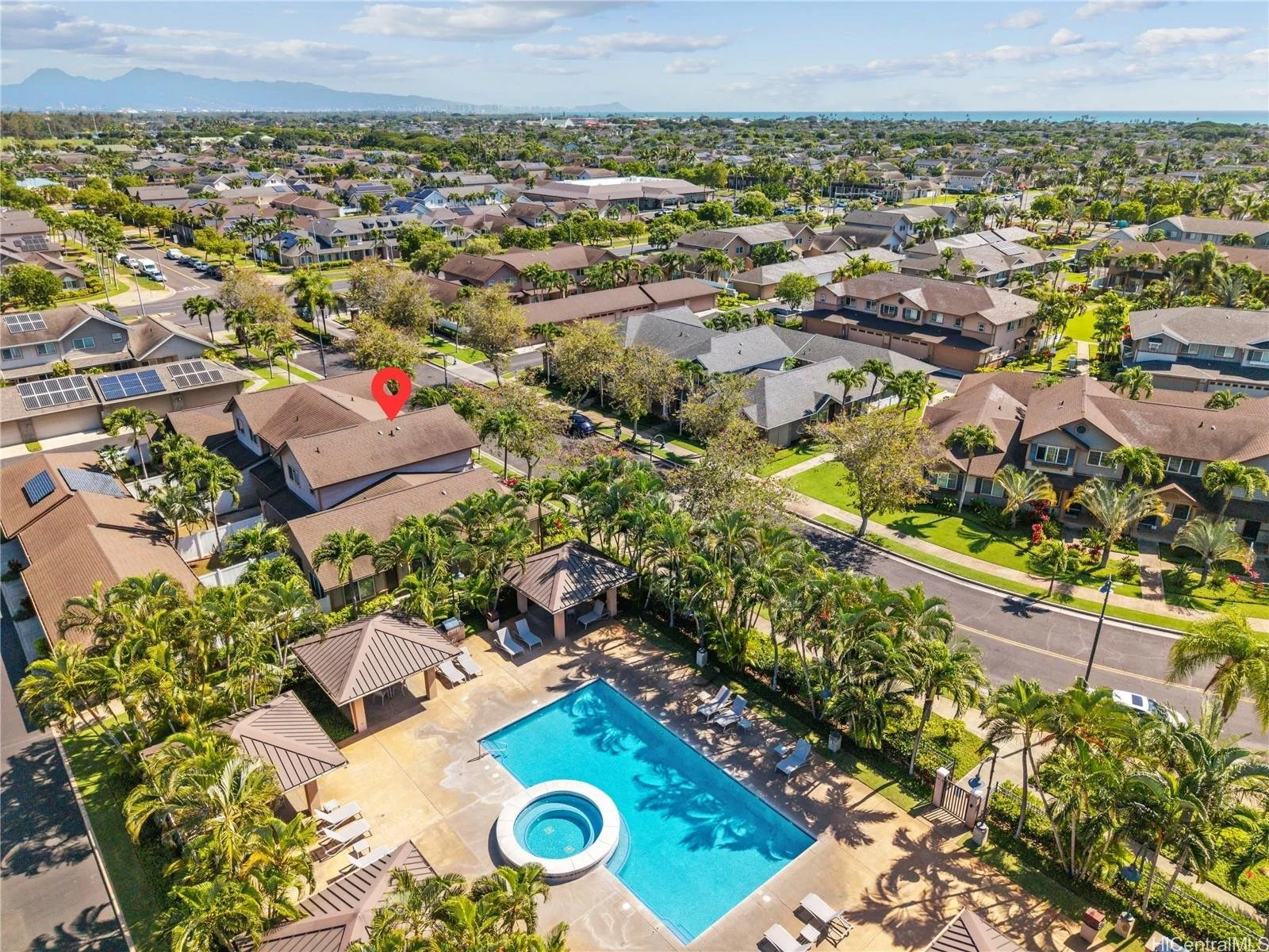 Aerial view of a residential neighborhood with houses, trees, and streets, featuring a community swimming pool in the foreground with umbrellas and lounge chairs.