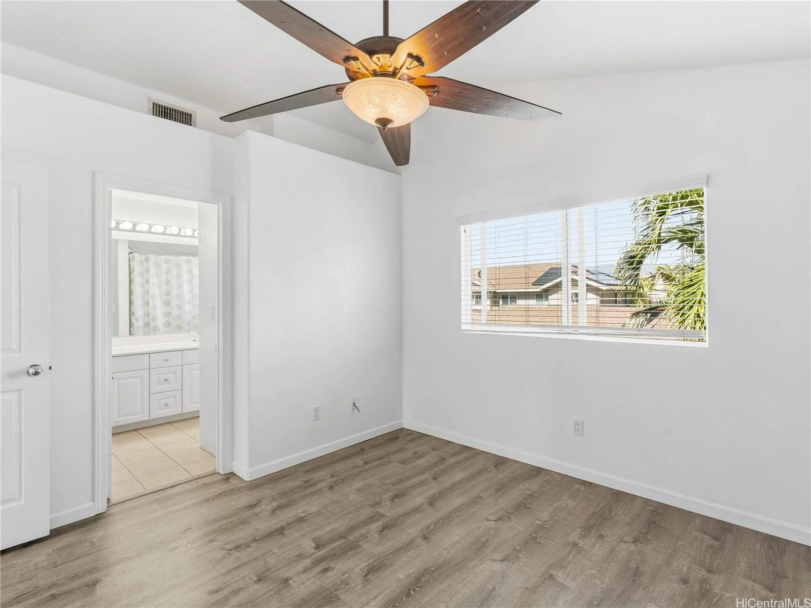 Empty bedroom with white walls, light wood flooring, a ceiling fan with wooden blades, and a window with blinds showing palm trees and neighboring houses.