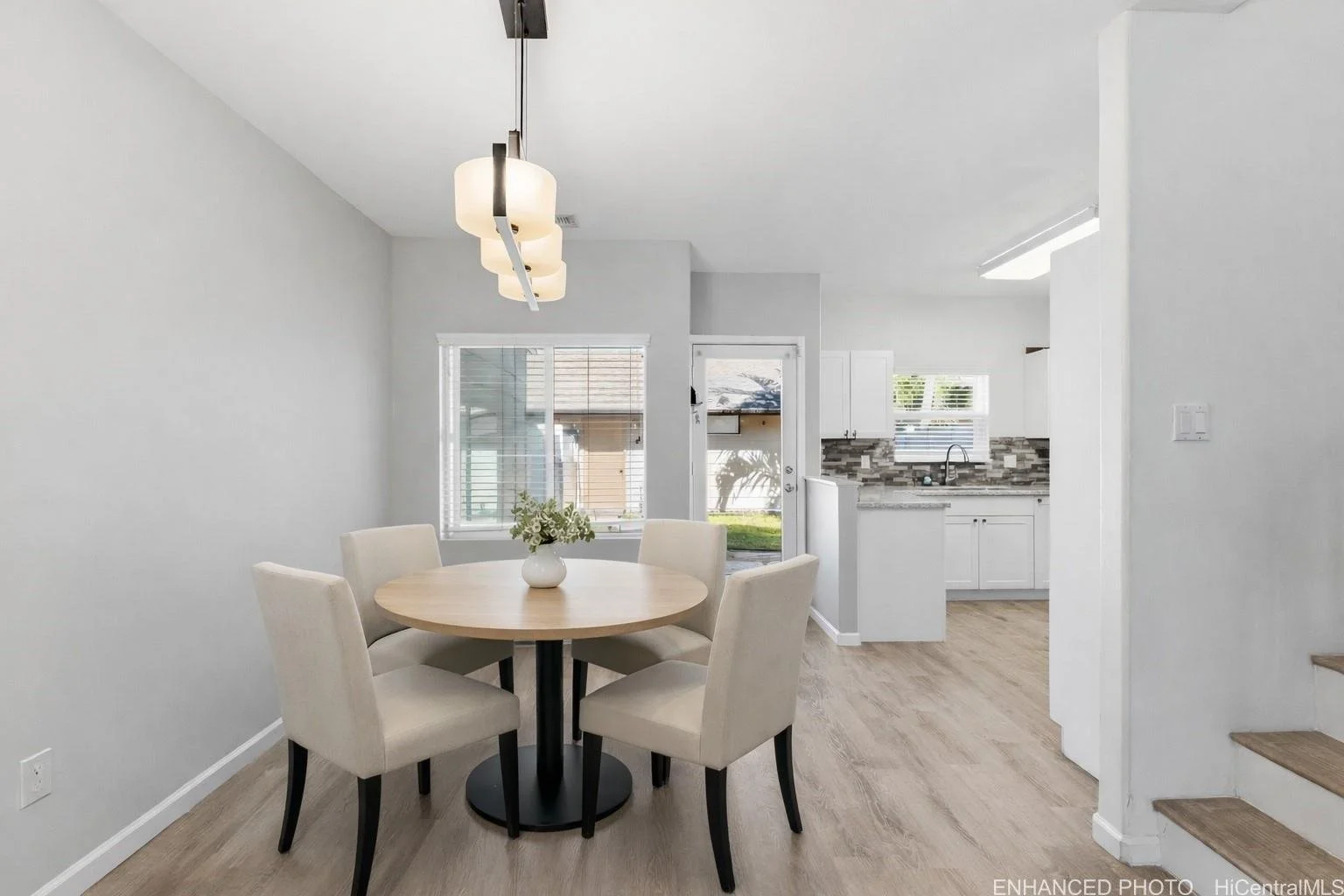 Dining area with a round table and six beige cushioned chairs, a vase with flowers, and a modern light fixture overhead. Visible kitchen with white cabinets, gray backsplash, and a window above the sink.