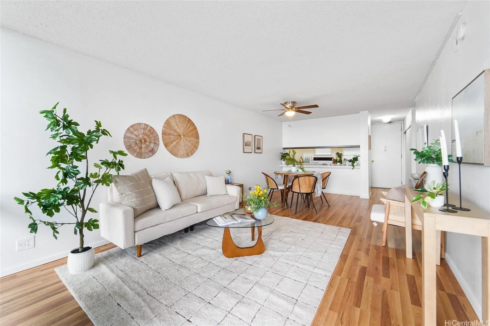 Living room with white sofa, wooden coffee table, green potted plant, beige rug, and wall decorations, connecting to a dining area with a round table and four chairs, wooden flooring throughout.