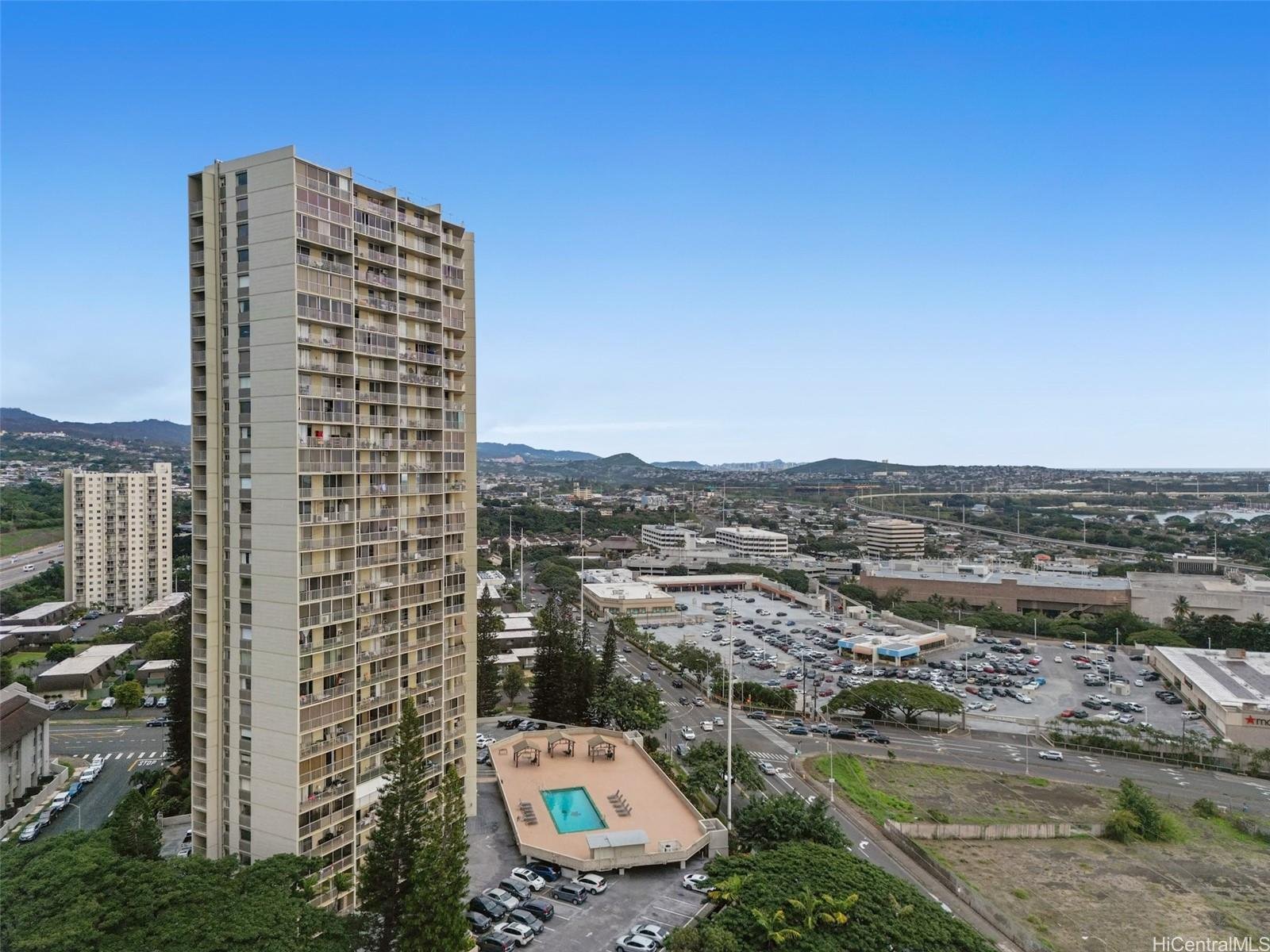 High-rise apartment building with balconies, a swimming pool and parking lot in the foreground, and a cityscape with roads and distant mountains under a clear blue sky.
