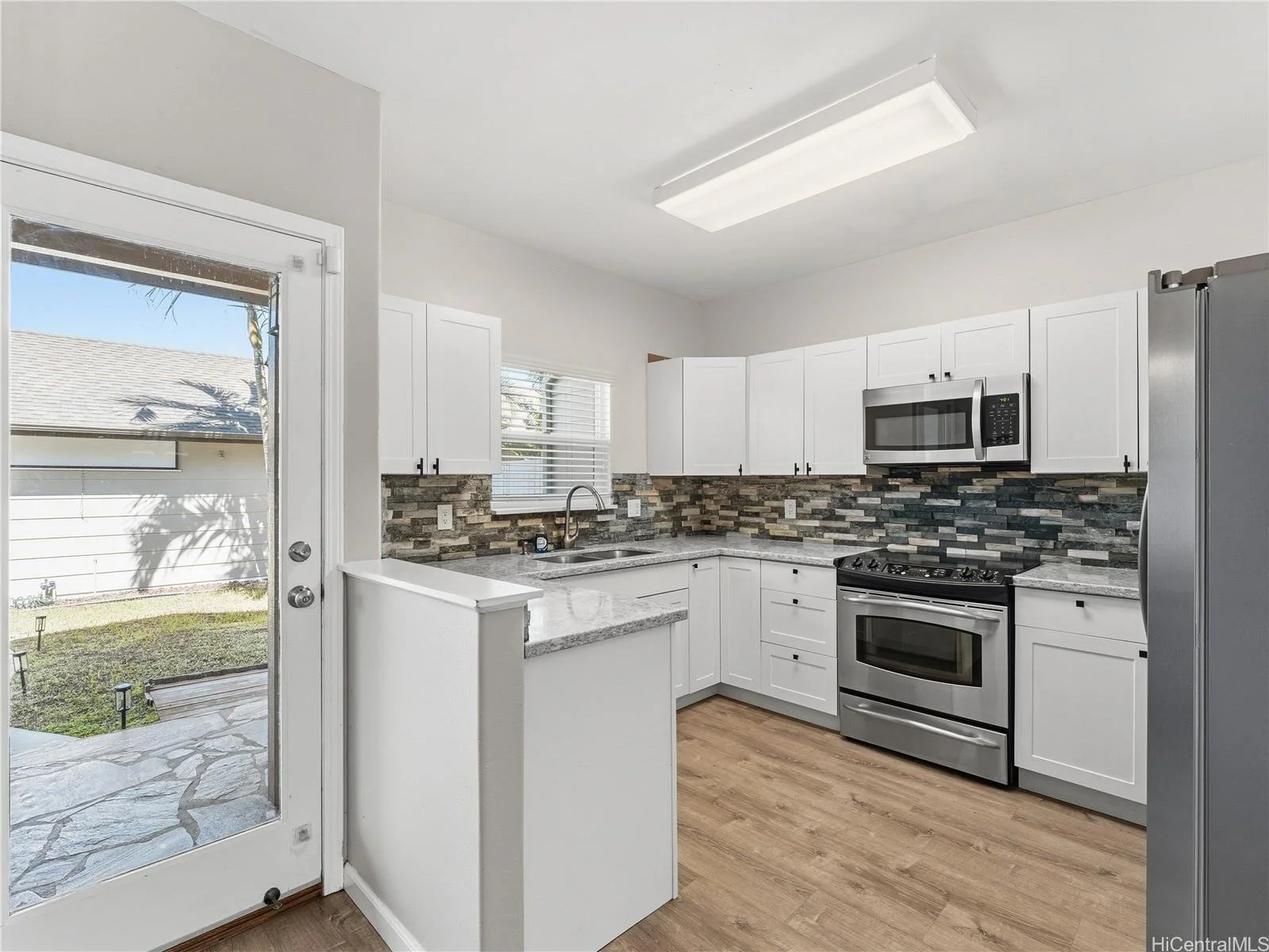 Kitchen with white cabinets, stainless steel appliances, a mosaic tile backsplash, and a door leading to a backyard with a stone patio.