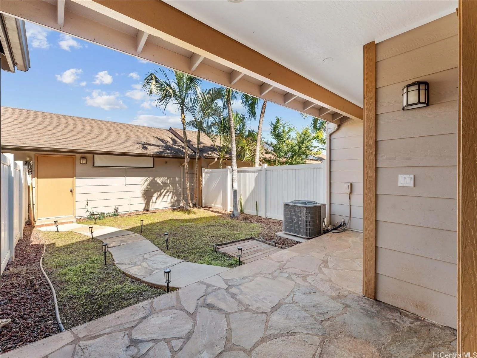 View of a backyard patio area with a stone pathway, small grass patches, and a white fence. Tall palm trees and a beige building with a sloped roof are visible. An air conditioning unit is placed on the patio.