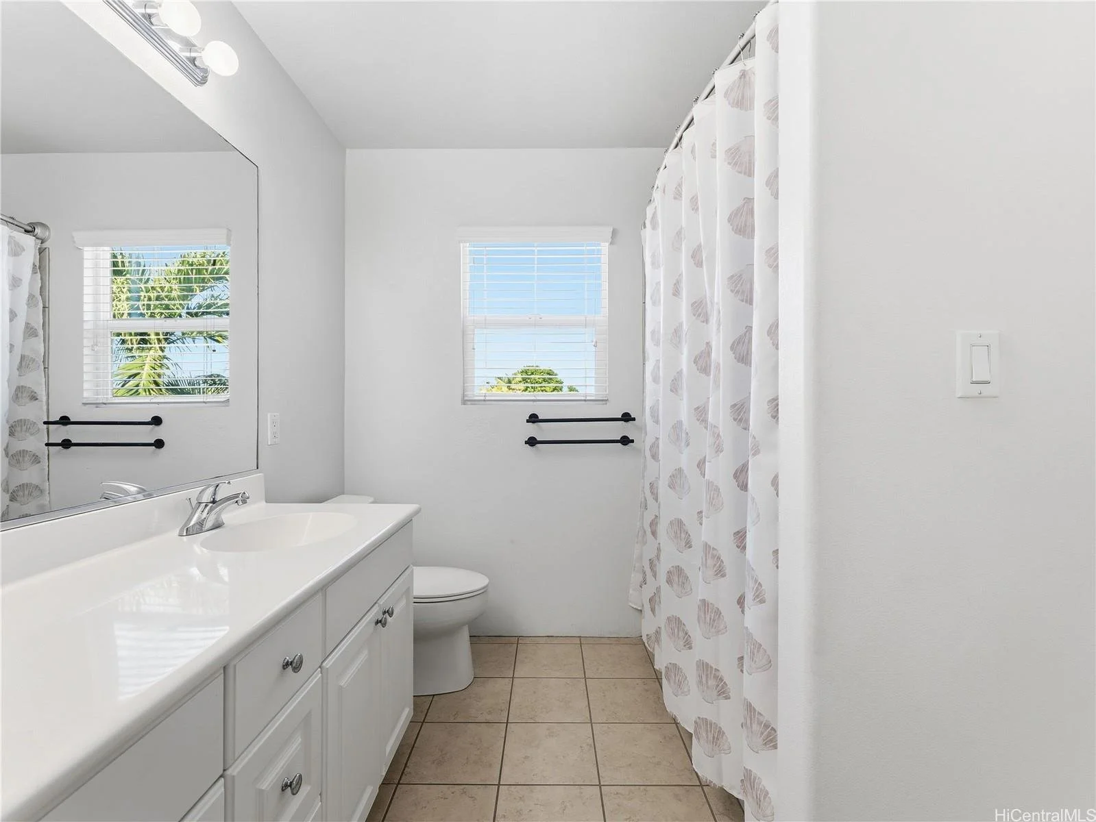 Bathroom with white vanity, large mirror, two windows, seashell-themed shower curtain, and beige tiled floor.
