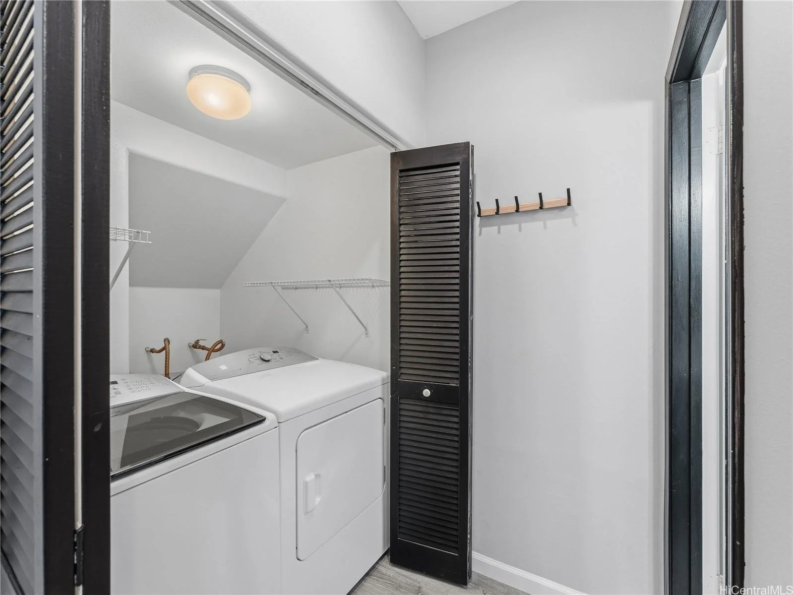 Laundry room with white washing machine and dryer, wall-mounted wire shelves, black louvered door, wooden coat rack, white walls, ceiling light, and gray flooring.