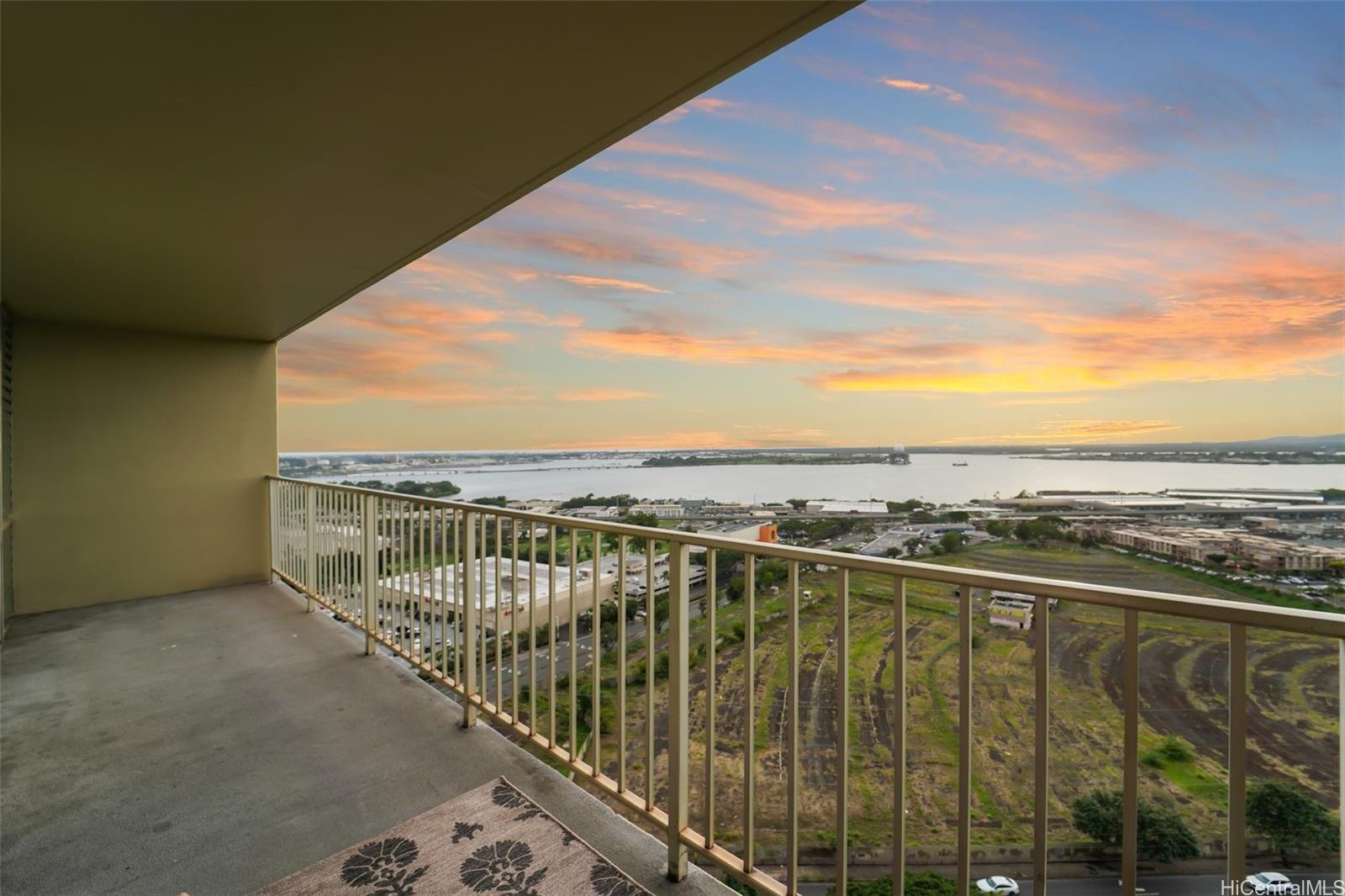View from a balcony over a river and surrounding landscape during sunset, with colorful sky and some buildings and fields visible.