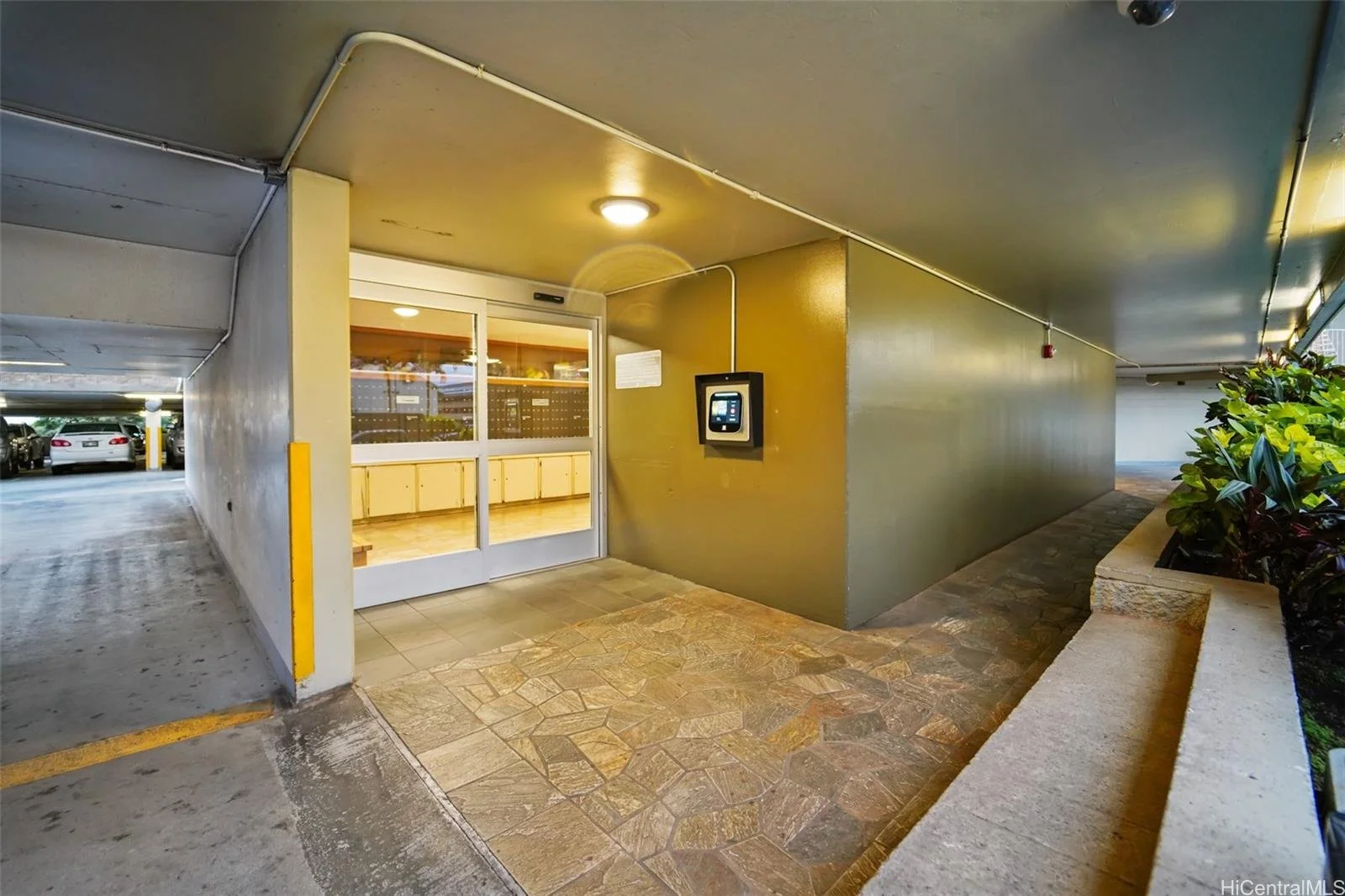 Entrance to a parking garage with a glass door, electronic access panel, and a small area with tiled flooring leading into a lobby area.