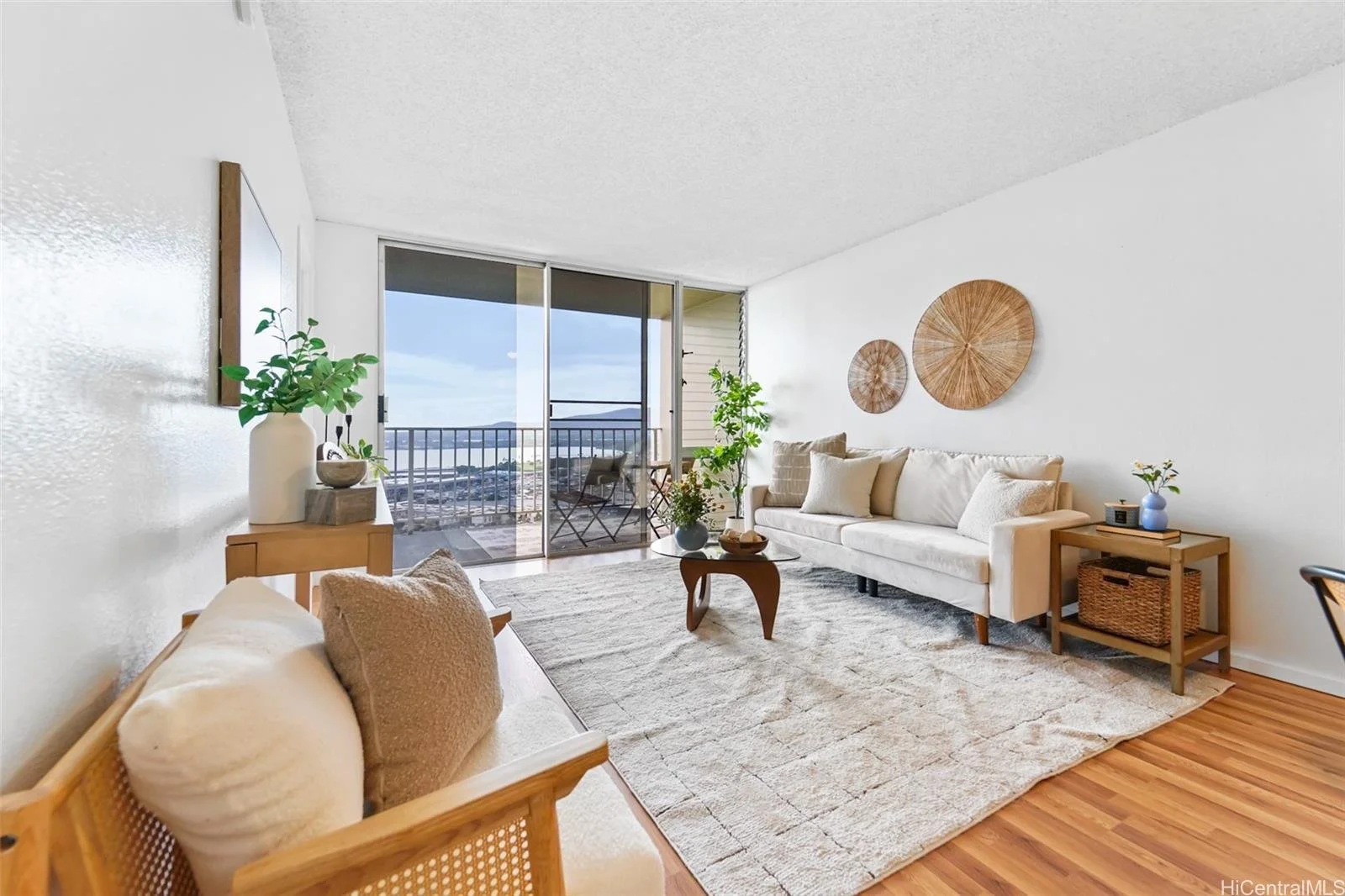 Bright living room with beige sofa, wooden side tables, potted plants, large sliding glass door leading to balcony with outdoor seating and ocean view, white walls decorated with wicker wall art, light-colored rug, wooden floors.