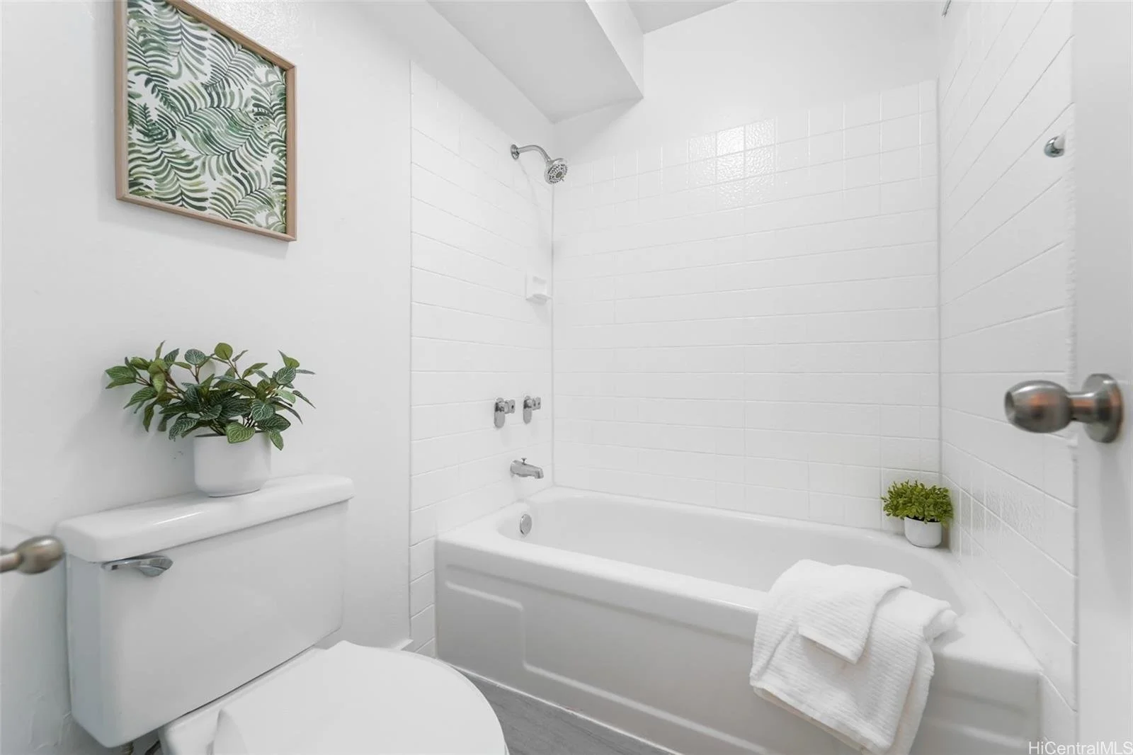 A clean, minimalist bathroom with white walls, a bathtub with a white towel draped over the edge, a small potted plant on the bathtub, a ladder shelf with a potted plant and framed artwork, and white tiled walls with a showerhead.