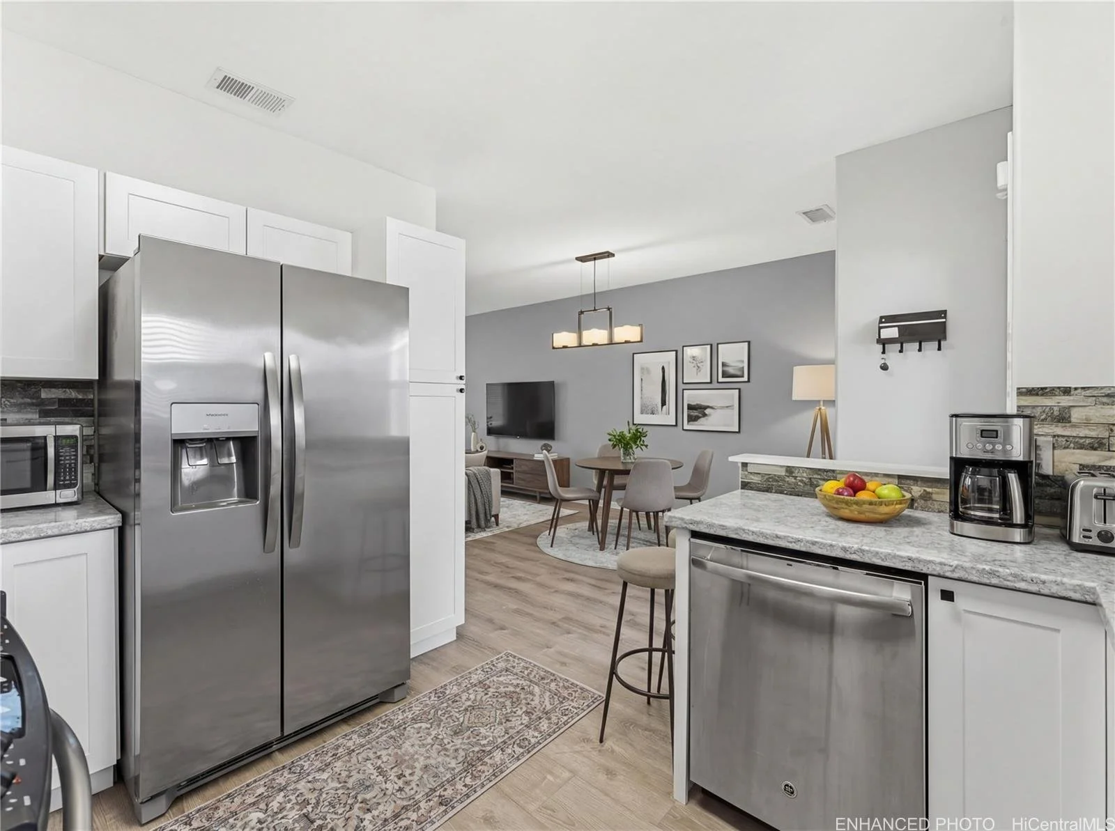 Modern kitchen with white cabinets, stainless steel refrigerator, microwave, coffee maker, toaster, and a bowl of fruit on the countertop, adjacent to a dining and living area.