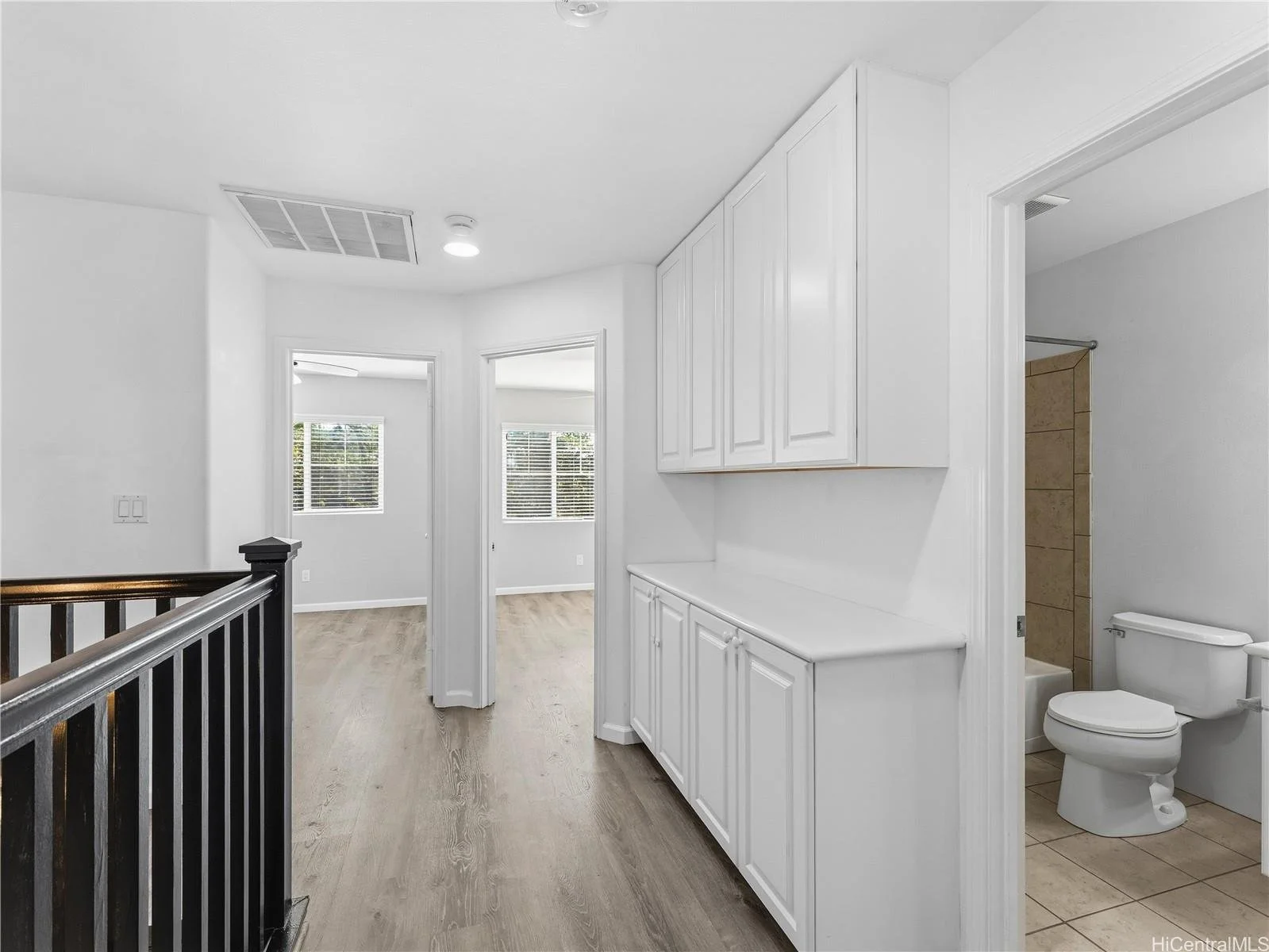 Interior of a modern house with white walls, wood flooring, a hallway leading to rooms with windows, a built-in white cabinet, and a bathroom with a toilet and shower visible.