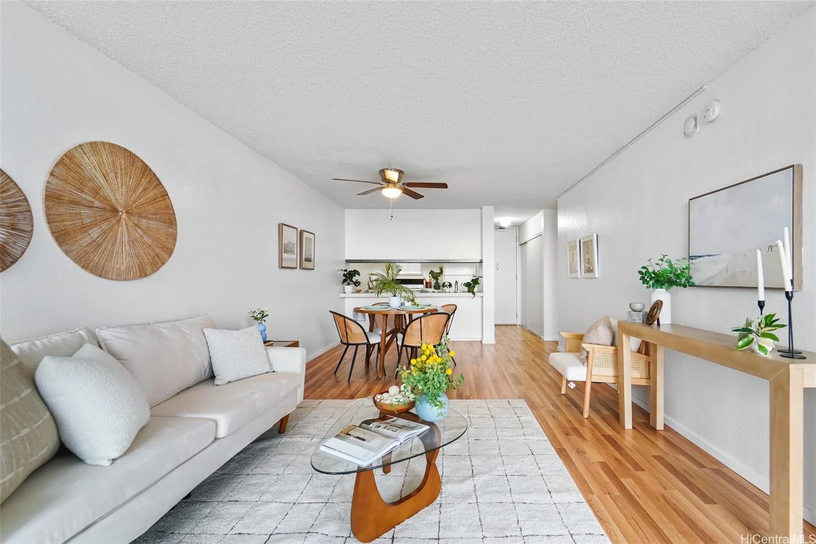 Bright living room with white walls, beige sofa with pillows, glass coffee table with a book and a vase, wooden flooring, dining area with a table and chairs, and decorative wall art.