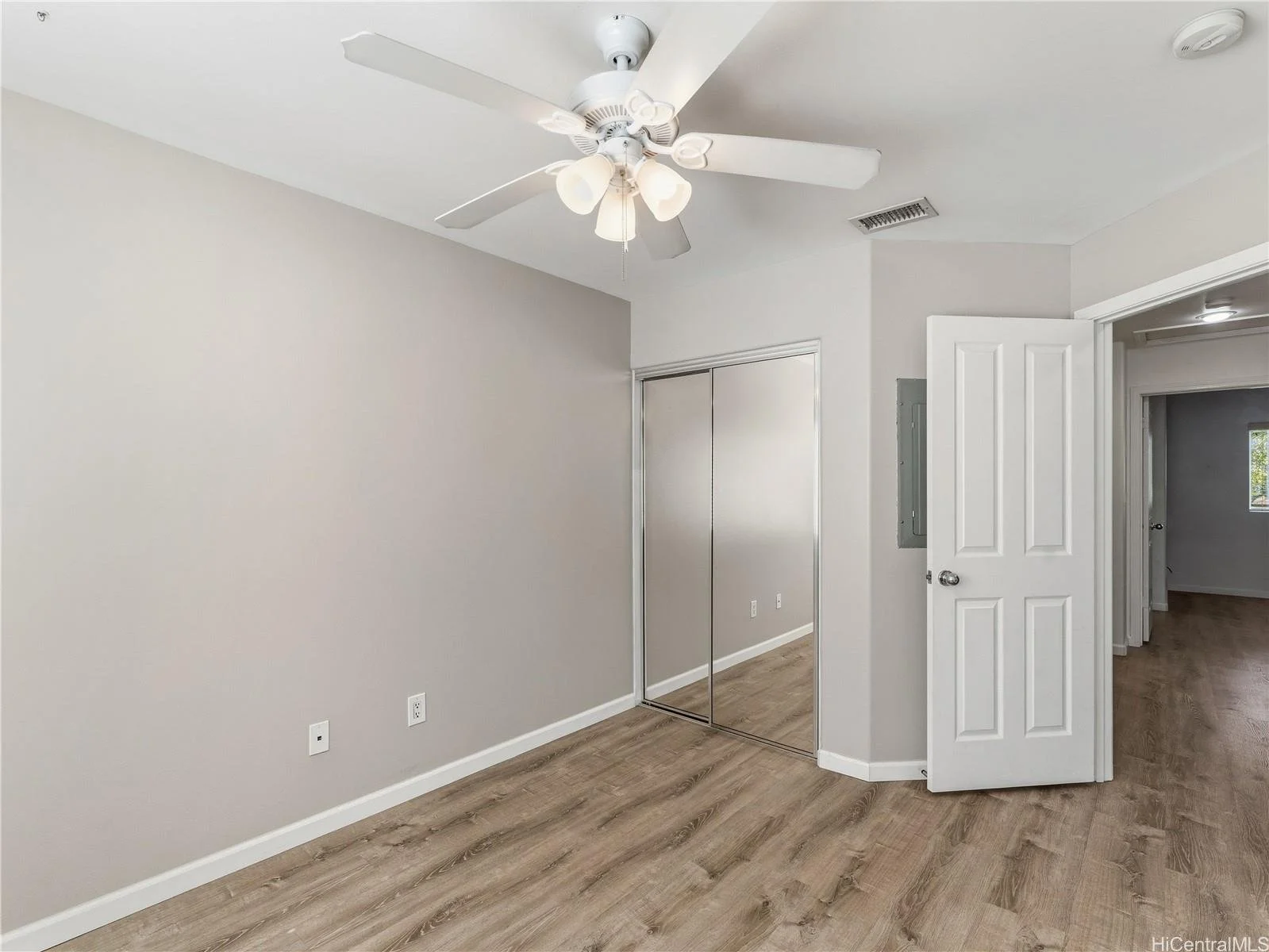 Empty bedroom with light-colored walls, wooden flooring, a ceiling fan with lights, a closet with mirrored sliding doors, and an open door leading to another room.