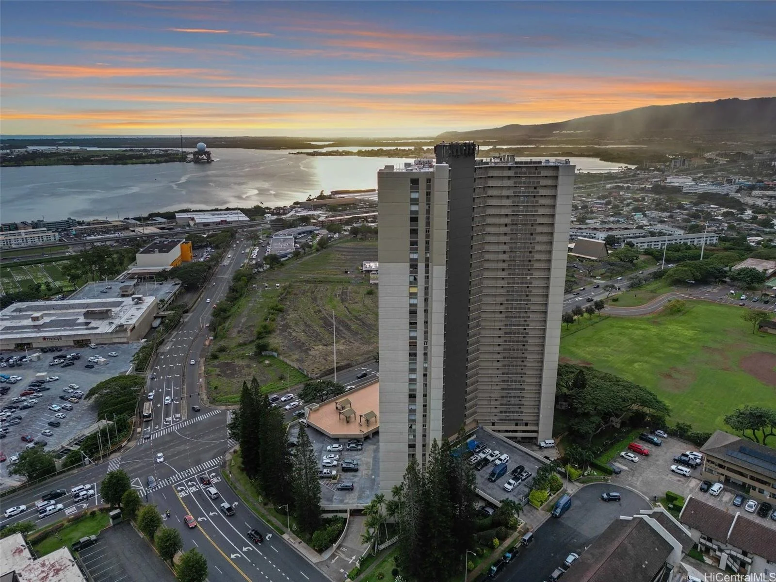 An aerial view of a cityscape at sunset, showing a water body, a tall apartment building, roads, parking lots, and green fields with a mountain in the background.