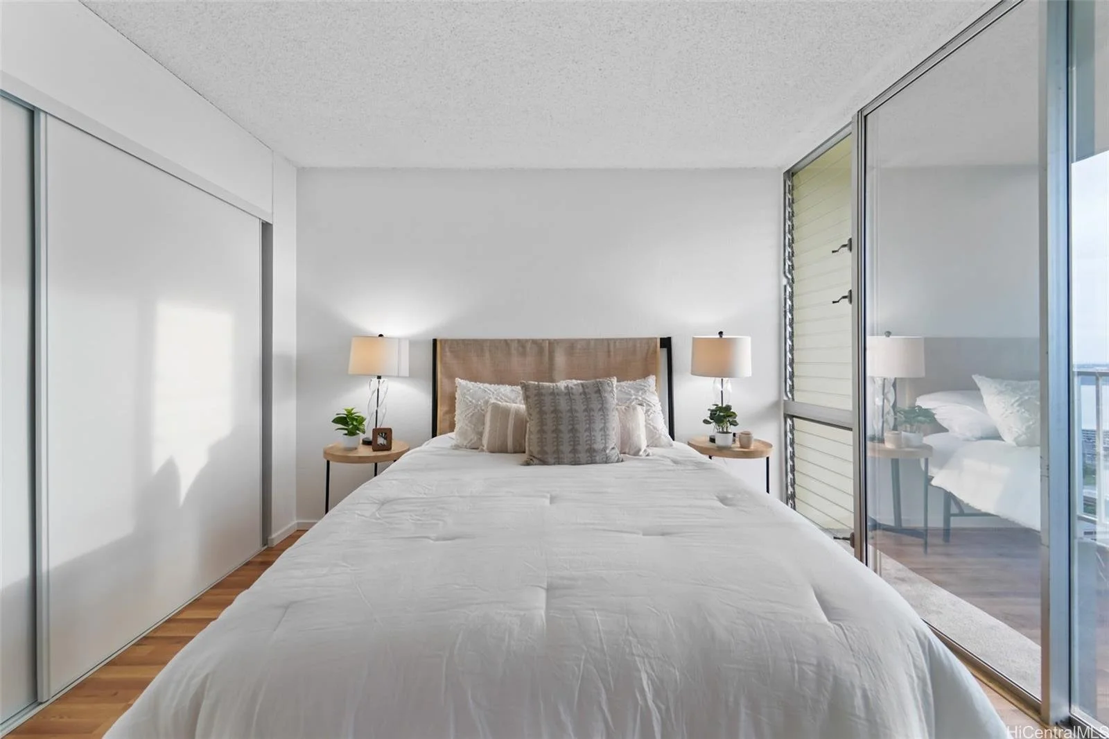 A bedroom with a bed, beige headboard, white bedding, multiple pillows, two bedside tables with lamps, and large windows with sliding glass doors leading to a balcony.