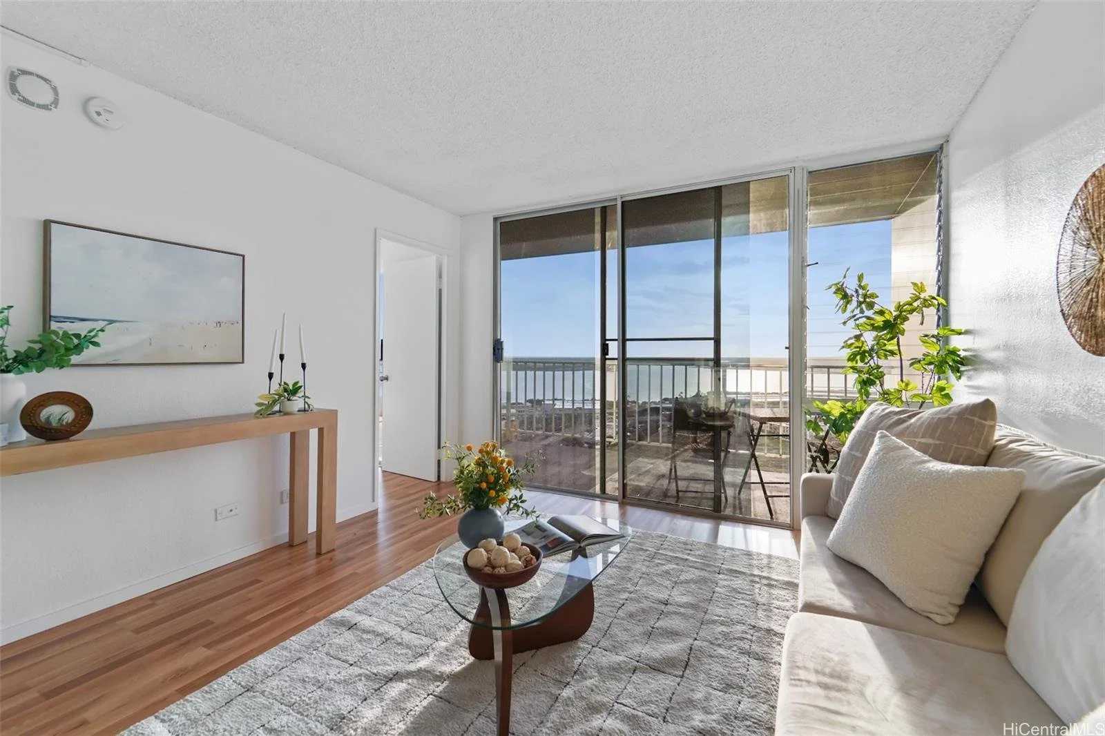 Living room with white walls, wooden flooring, a beige sofa with pillows, a glass coffee table with flowers, a large sliding glass door leading to an outdoor balcony, and a decorative wall hanging.