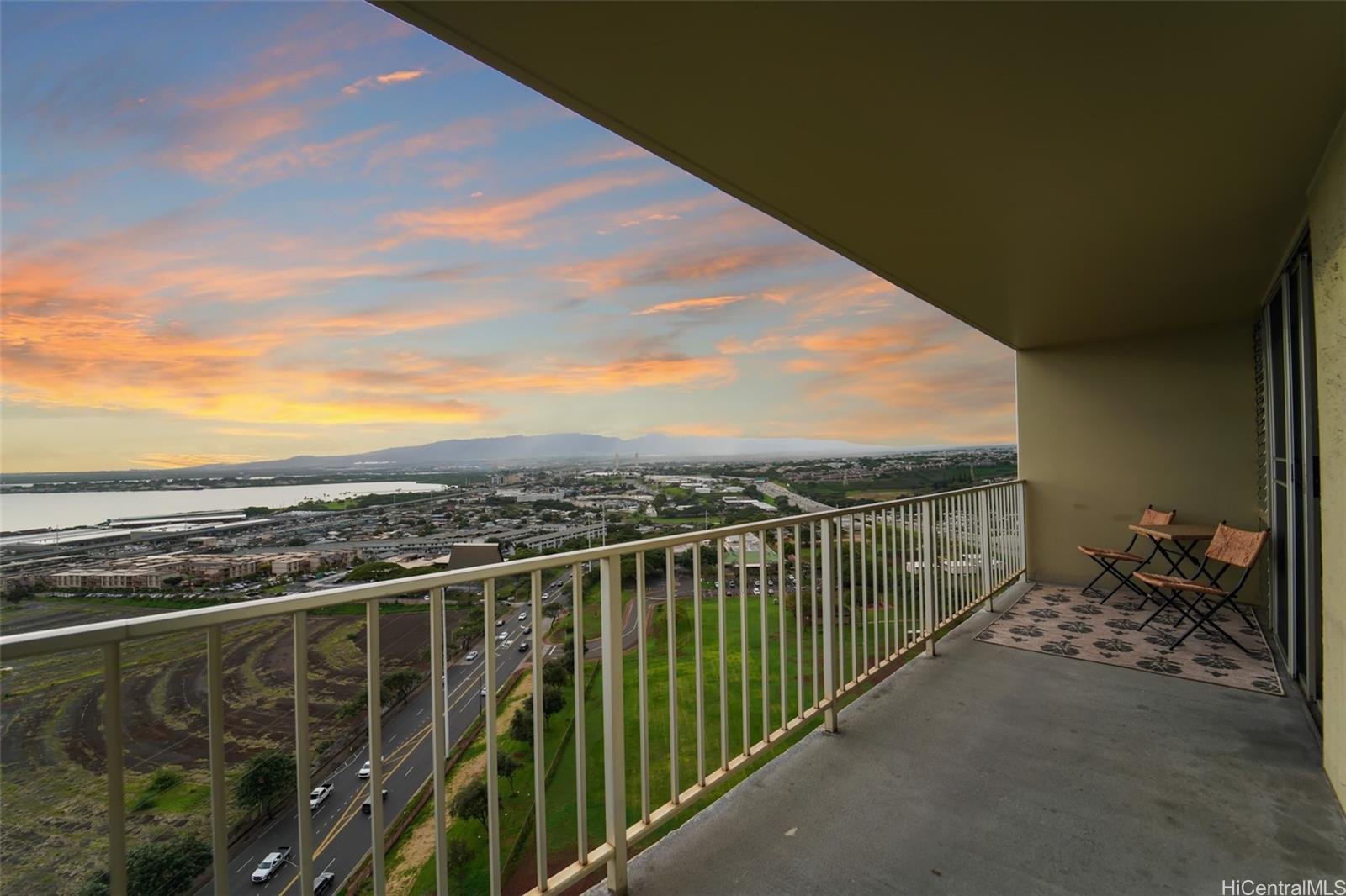 View from a high-rise balcony overlooking a city skyline, river, and mountains at sunset, with two chairs and a small table on the balcony.