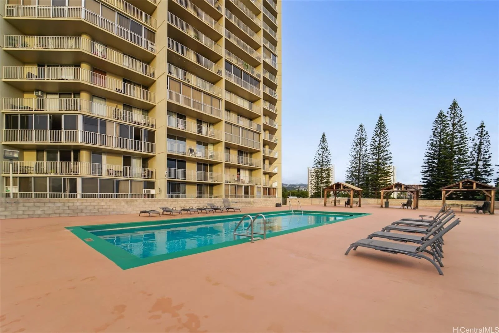Apartment complex pool area with lounge chairs, small cabanas, and a high-rise building in the background, surrounded by trees.
