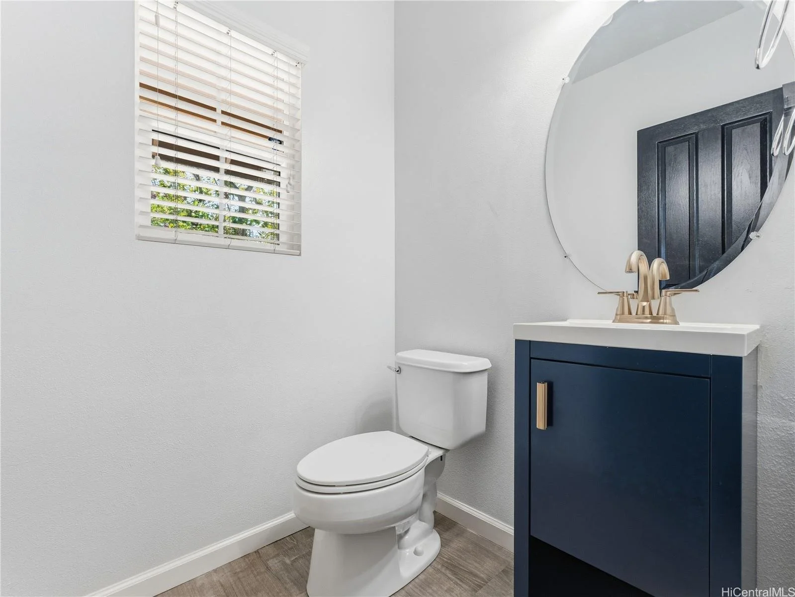 Small bathroom with a white toilet, a navy blue vanity with a white countertop, a round mirror, a gold faucet, and a window with white blinds.