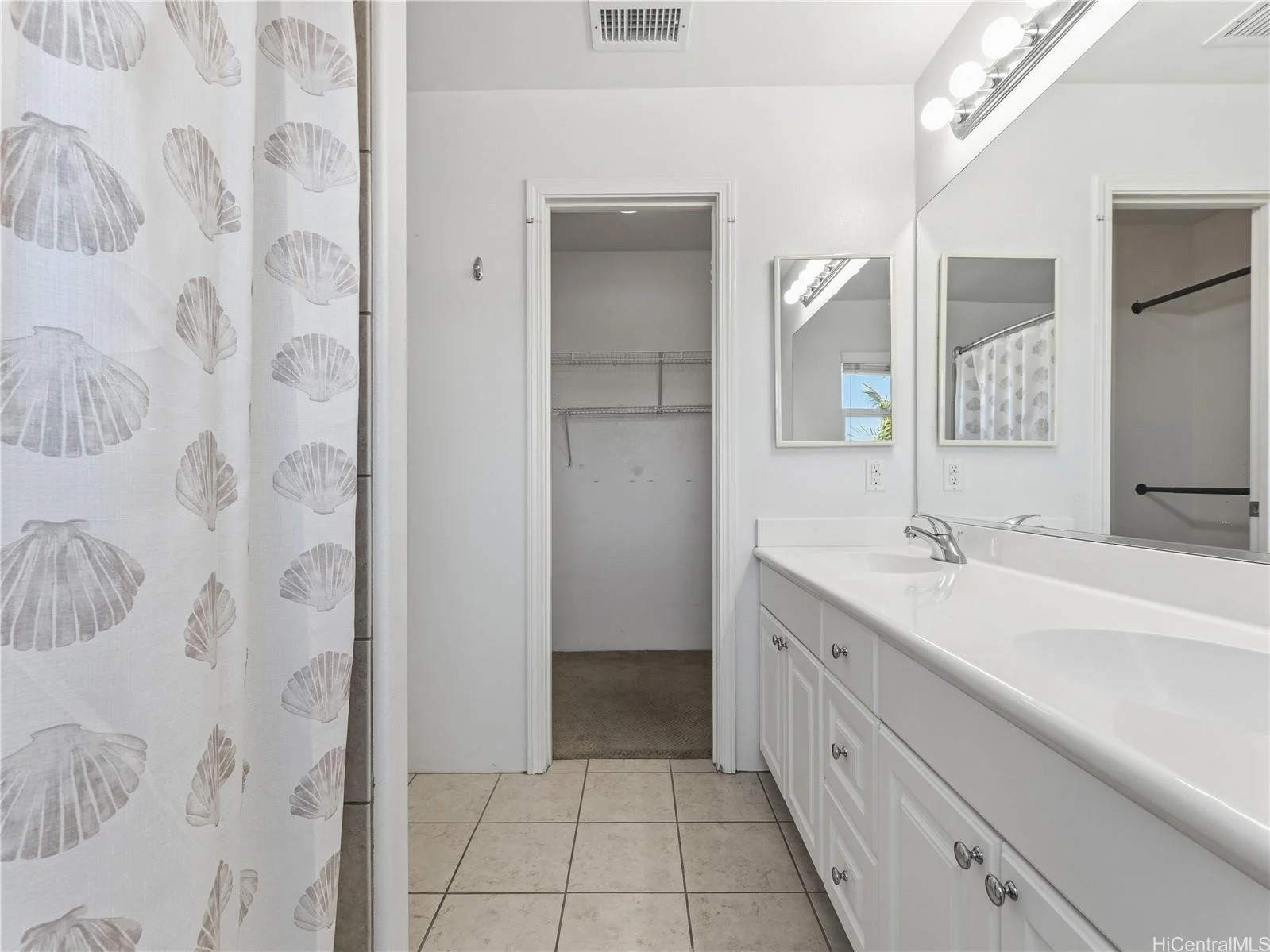 A bathroom with a white vanity, large mirror, two small mirrors, and a shower with a shell-patterned curtain. An open closet space is seen with wire shelves. The bathroom has beige tiled flooring and white walls.