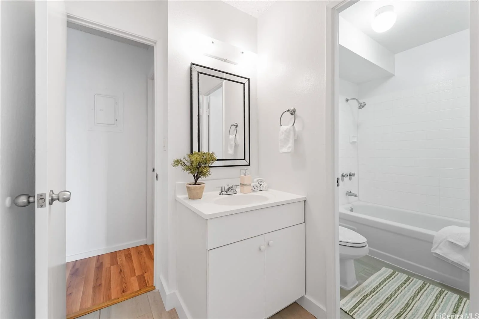 A small bathroom with white walls, a white vanity with a mirror, a potted plant, and rolled towels. A separate room with a bathtub and shower has a striped rug on the floor.