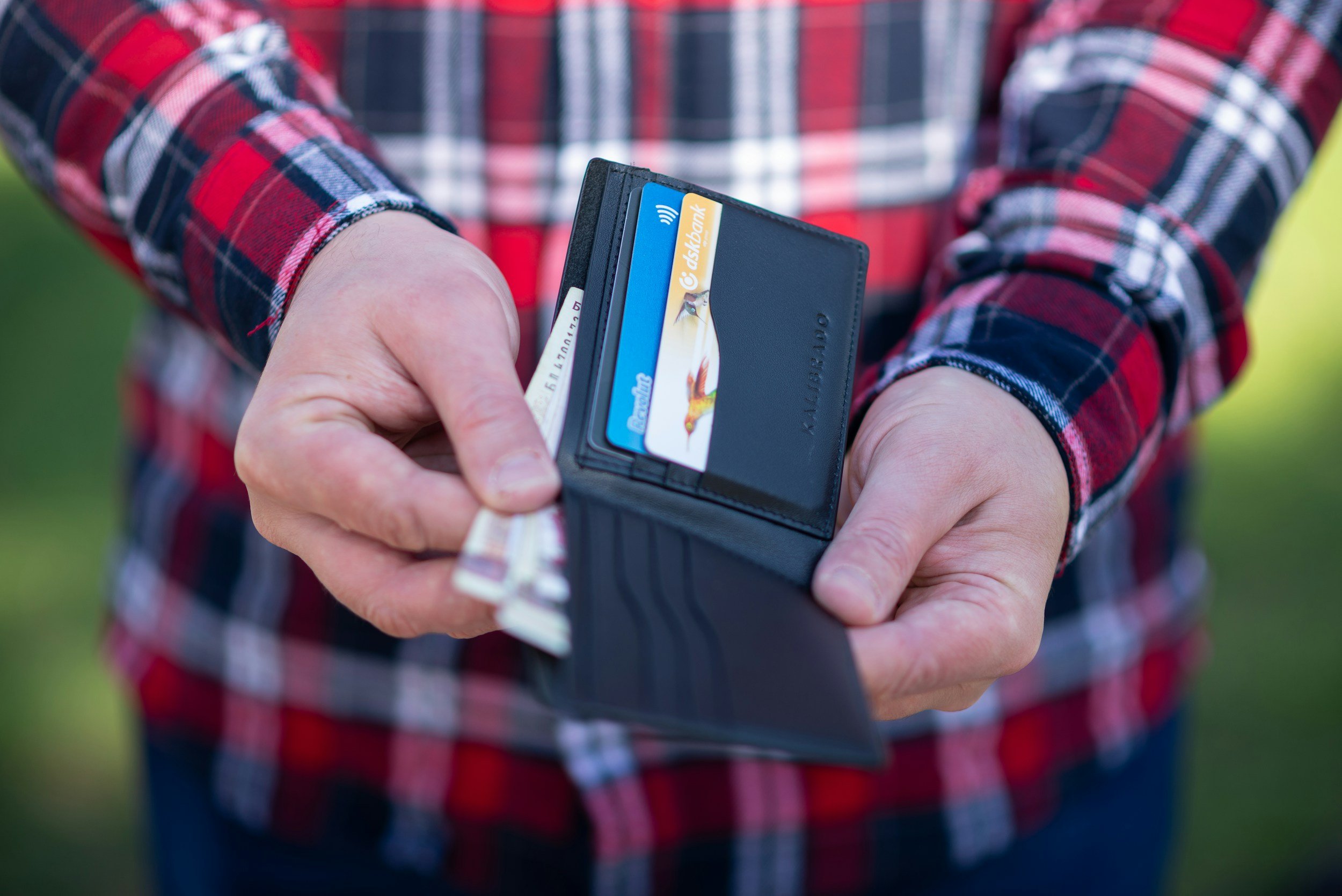 Person holding a wallet with credit cards, cash, and a bank card inside, wearing a red plaid shirt.