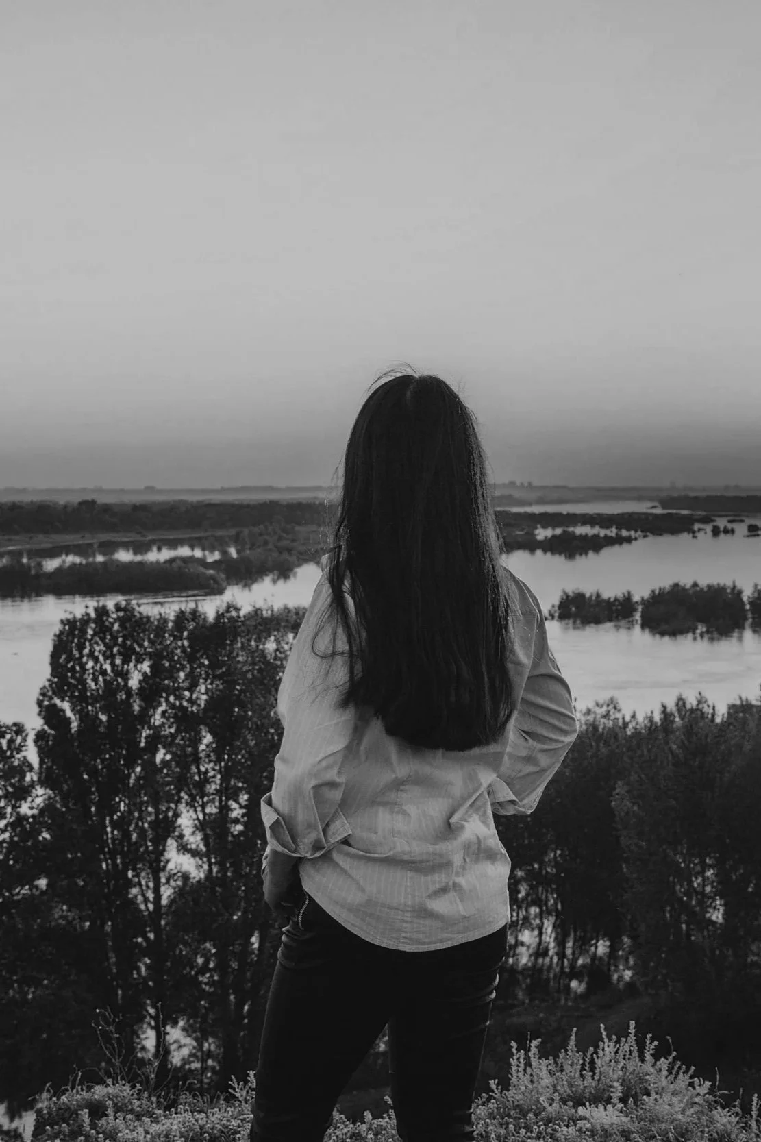 A woman with long dark hair, wearing a light-colored jacket and dark pants, stands with her back to the camera and gazes over a river landscape with trees and distant land, in black and white.