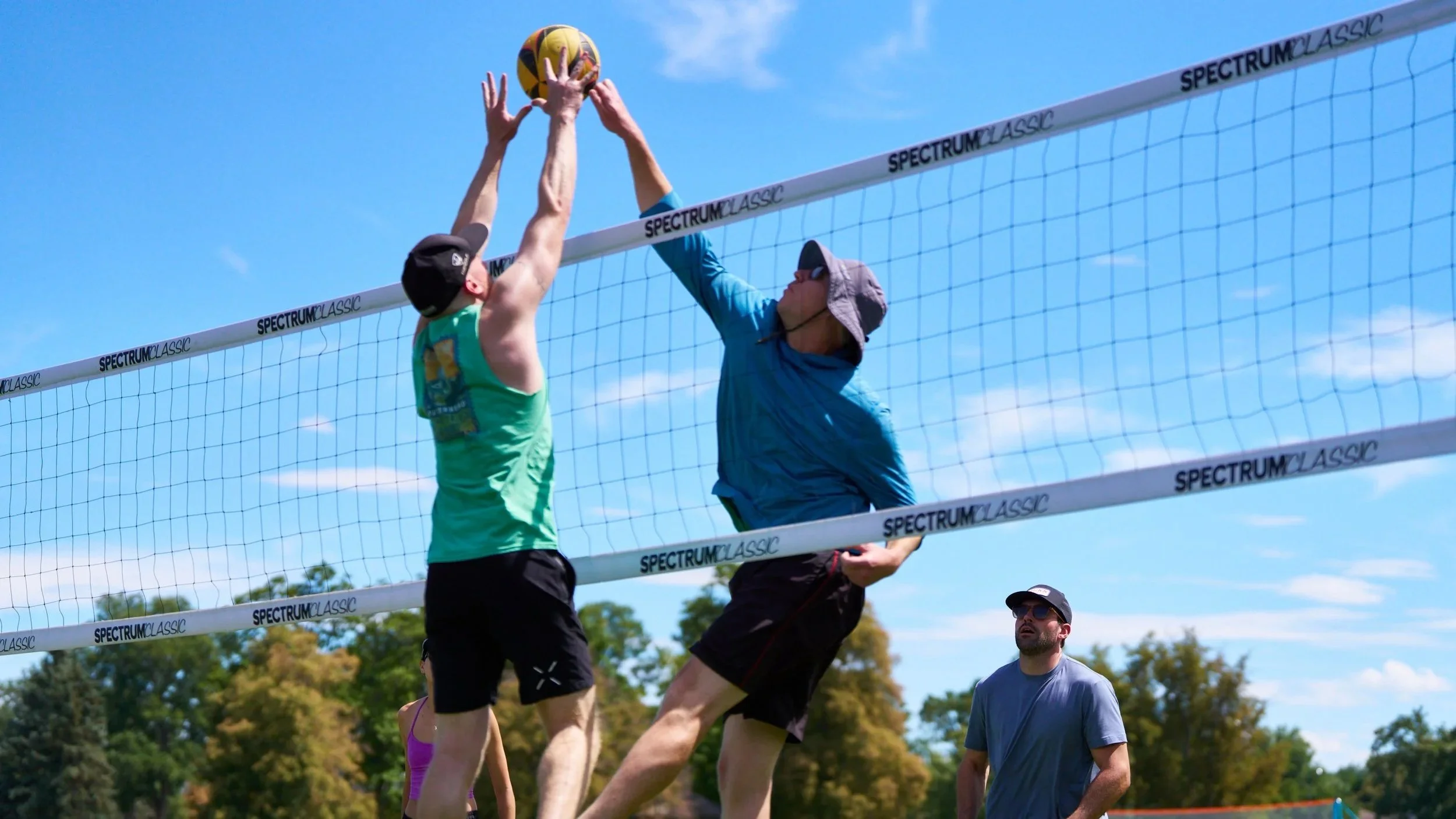 Two male volleyball players jumping to hit the ball over the net, with a third man watching nearby on a sunny outdoor court.