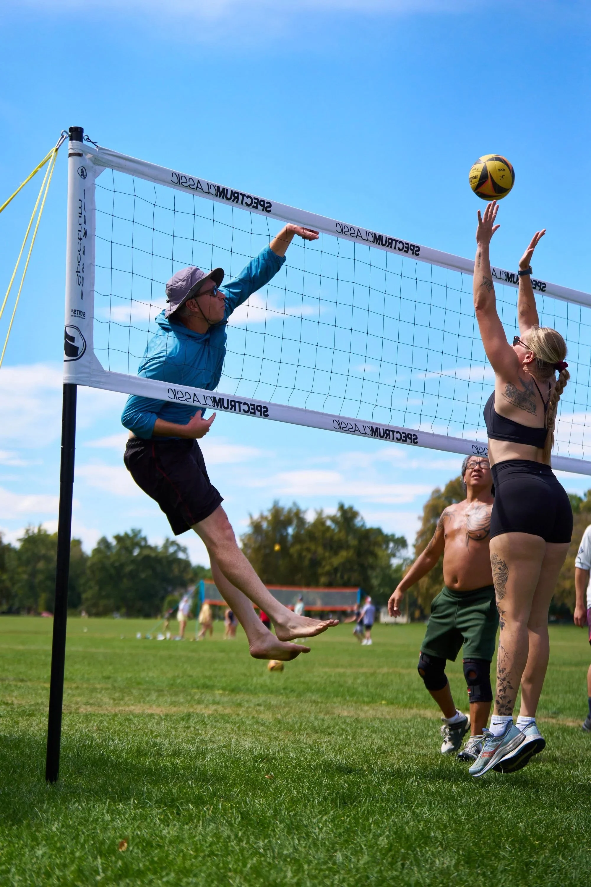 A volleyball game is taking place outdoors on a grassy field. Two women are jumping up near the net, with one hitting the ball and the other attempting to block. Other people are visible in the background, and trees and a blue sky are overhead.