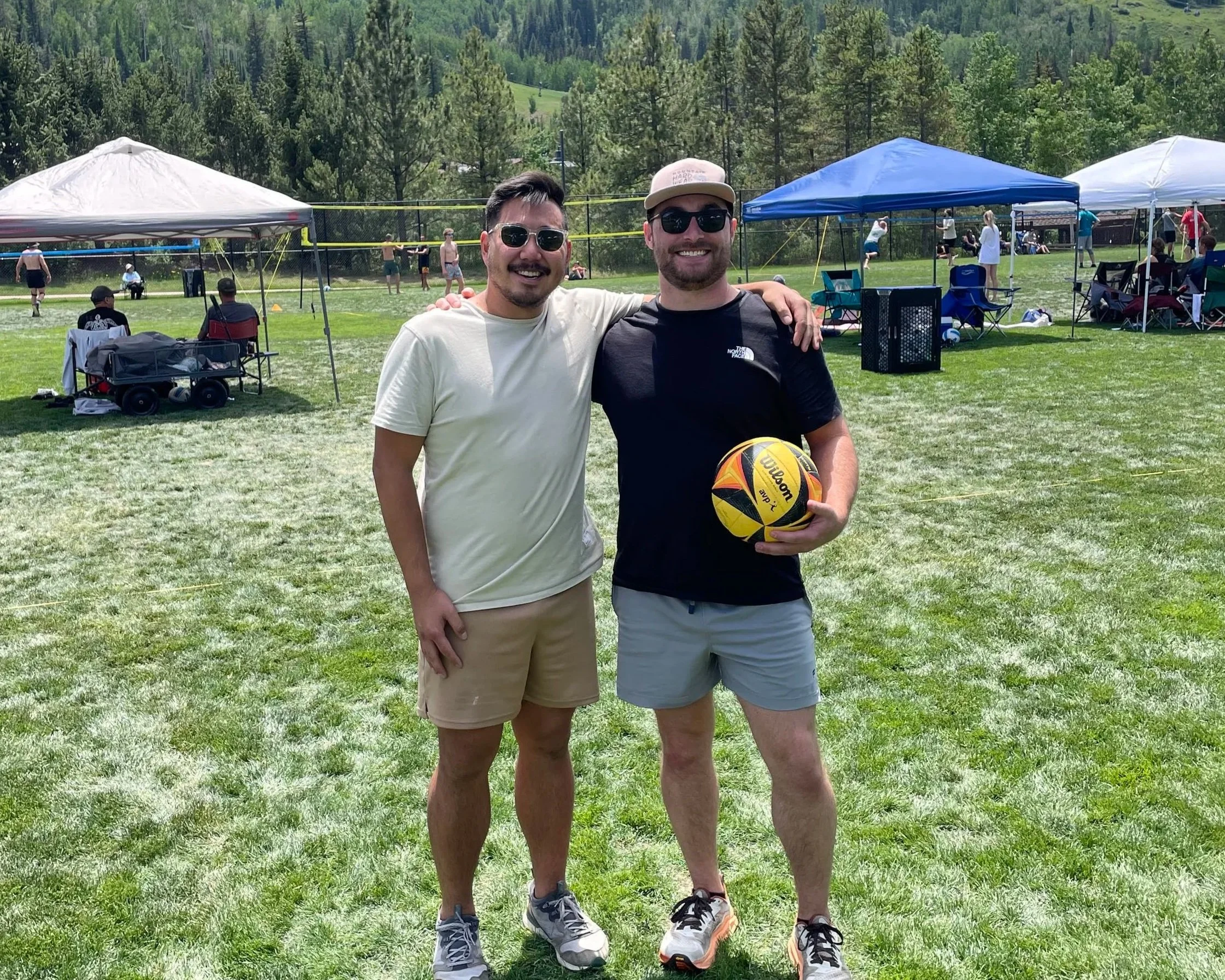 Two men smiling, standing on grass at an outdoor recreational event, with tents, volleyball net, and people playing volleyball in the background, surrounded by trees and mountains.