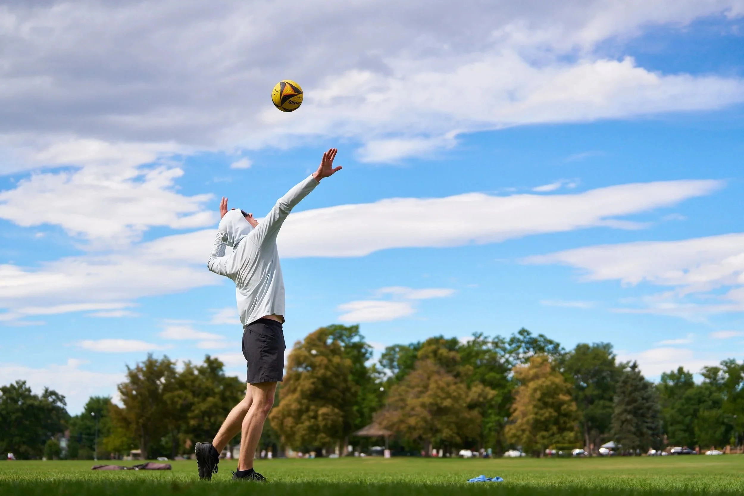 A person playing volleyball outdoors, jumping to hit the ball in a grassy park with trees and a partly cloudy sky.