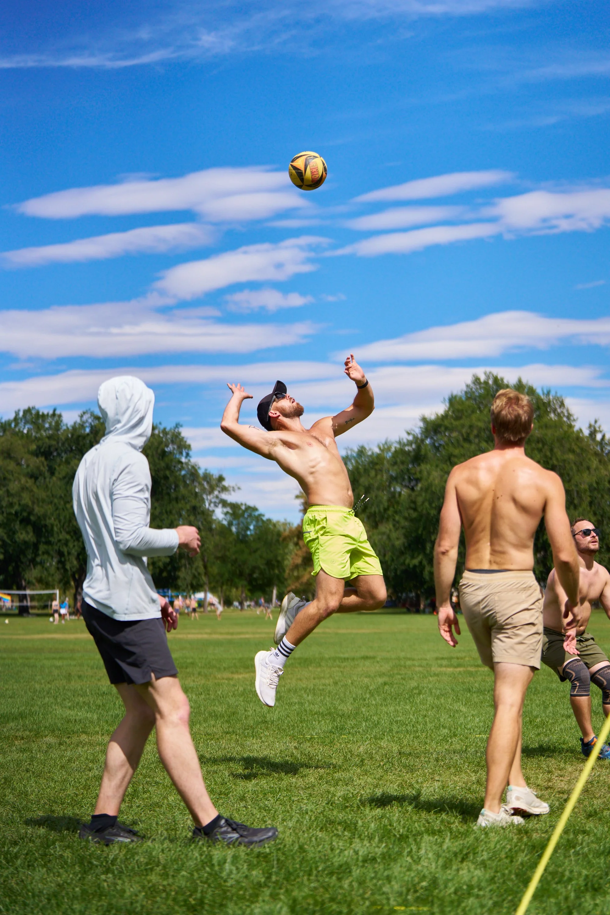 Young man in yellow shorts jumping to spike a yellow and black ball during a game on a grassy field, with other players under a blue sky with white clouds and trees in the background.