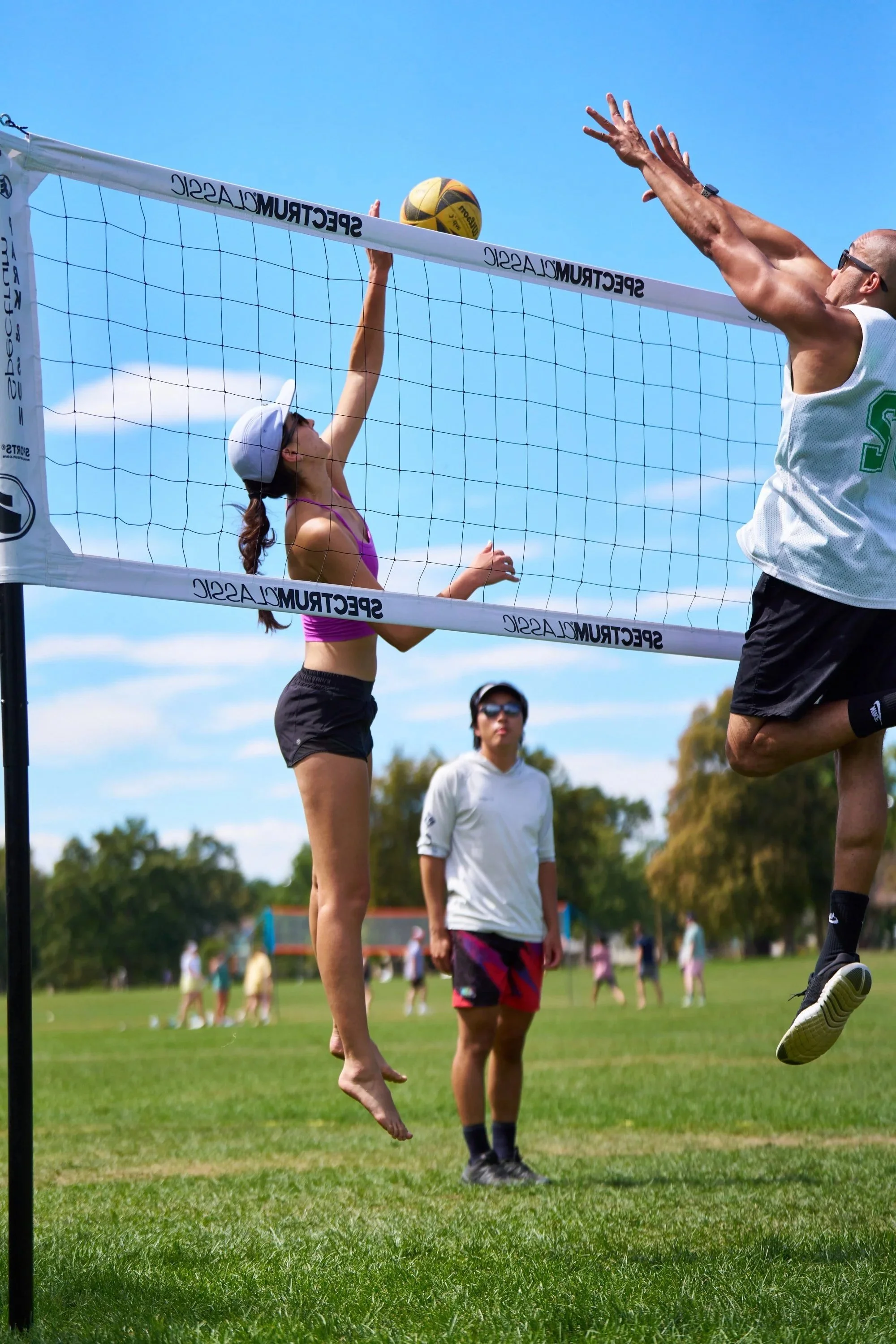Two people playing volleyball outdoors on a sunny day; the woman is jumping to hit the volleyball over the net, and the man is jumping to block the ball.