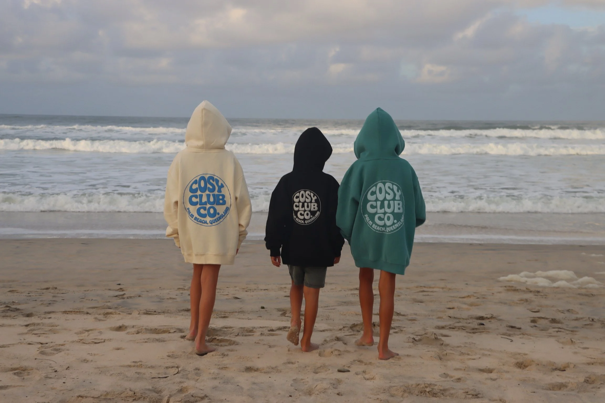 Three children, two girls and one boy, walking along the beach, wearing hooded sweatshirts with 'COSY CLUB CO' logos on the back, towards the ocean with waves and a cloudy sky.