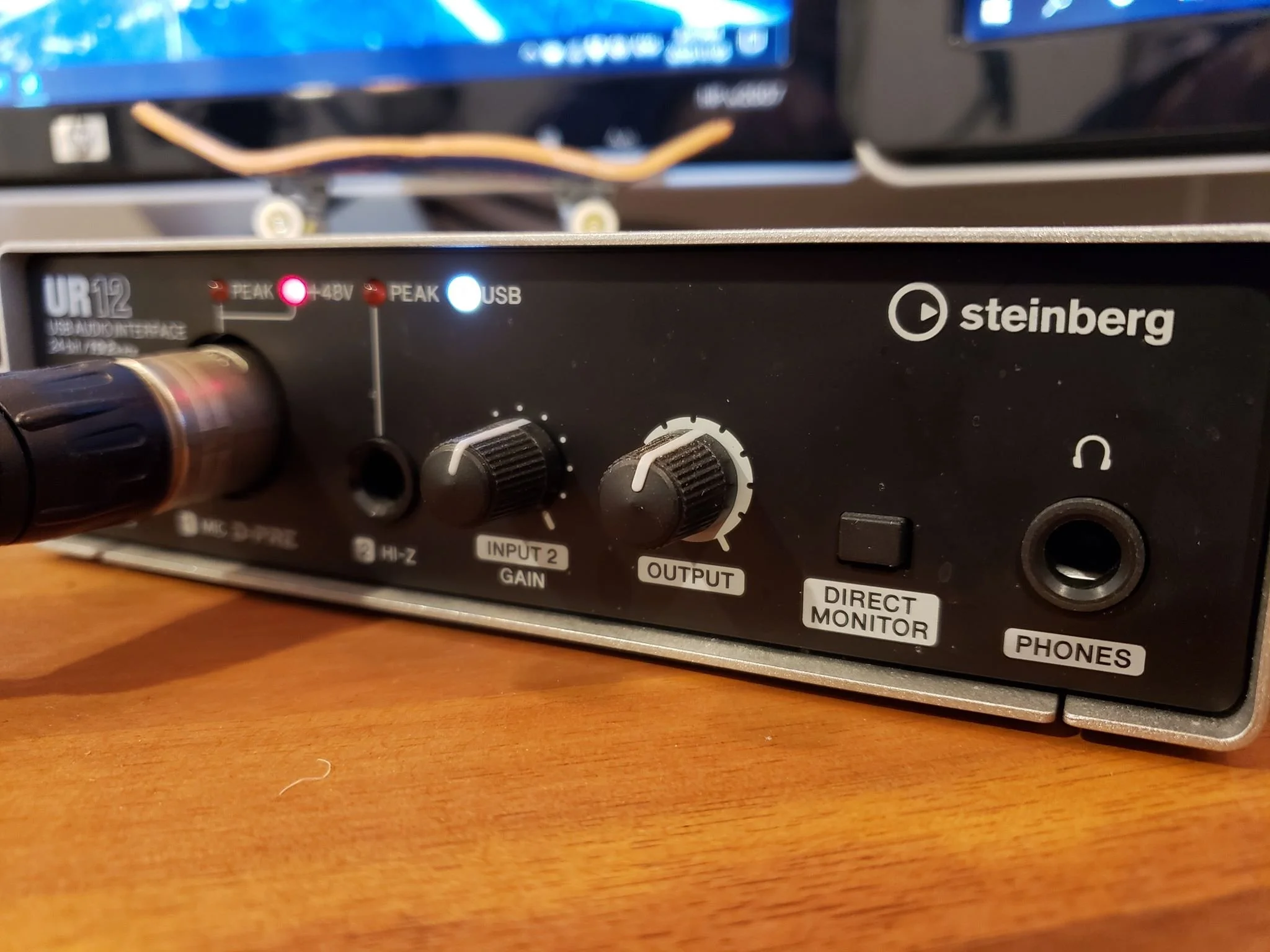 Close-up of a black Steinberg UR22 audio interface with various knobs, ports, and indicator lights, resting on a wooden surface. The interface has a USB port, gain and output controls, and labeled connections for headphones and direct monitor.