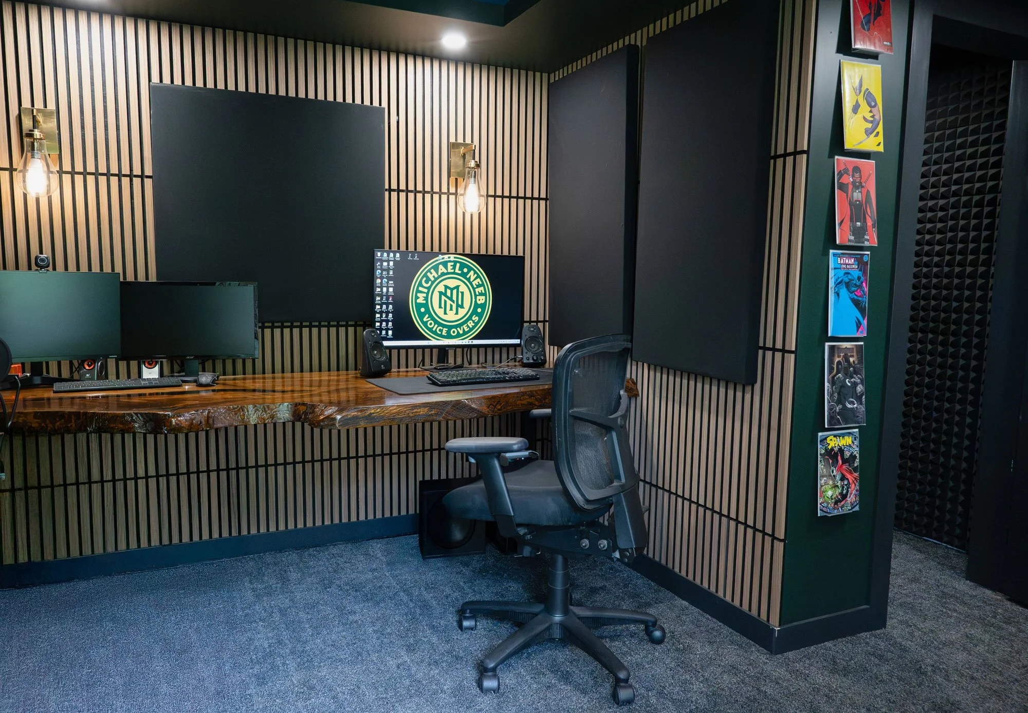 An office space with a wooden desk holding dual computer monitors, a keyboard, a mouse, and two speakers. Behind the desk is a black mesh office chair. The wall features wood paneling with a large black sound panel and two vintage-style light fixtures. To the right, a vertical display of comic book covers is mounted on the wall.