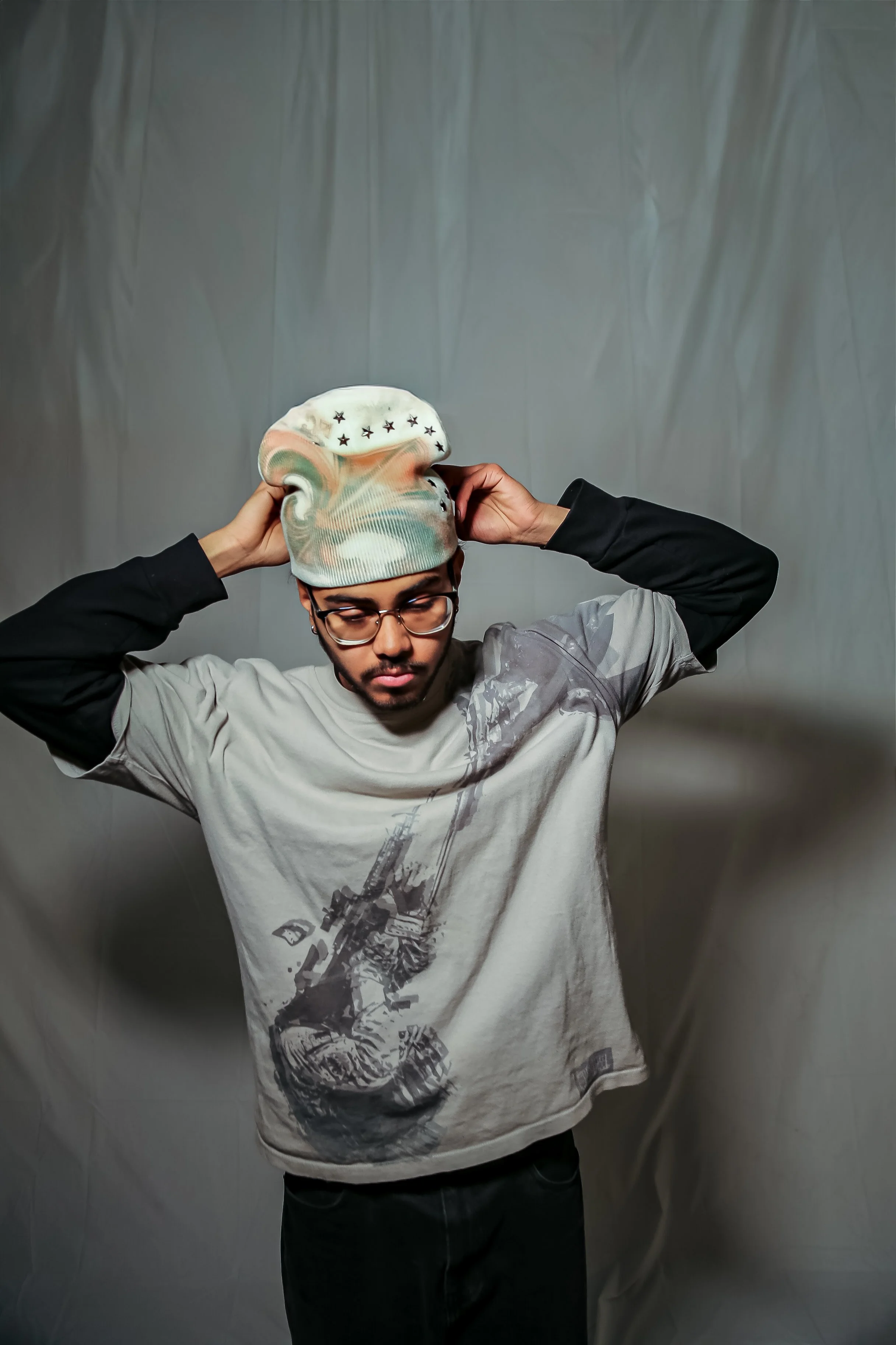 A young man wearing glasses, a gray graphic t-shirt featuring a construction machine, and a cap with star patterns, is adjusting his cap with both hands against a plain gray background.