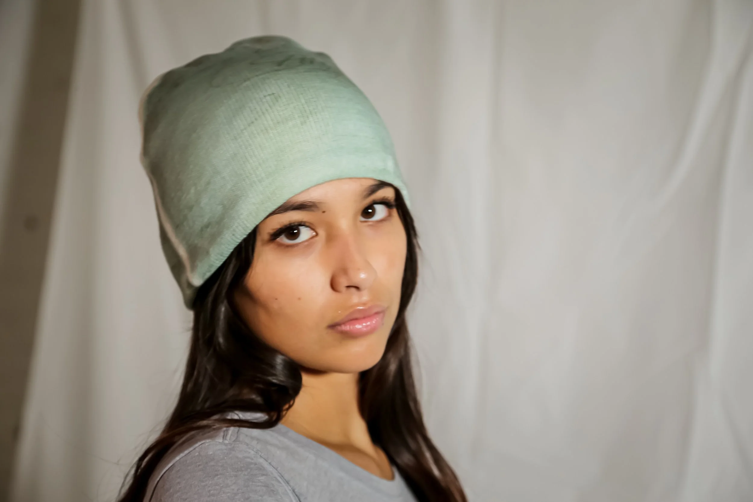 Young woman with long dark hair wearing a light gray shirt and a green beanie, looking at the camera against a plain background.