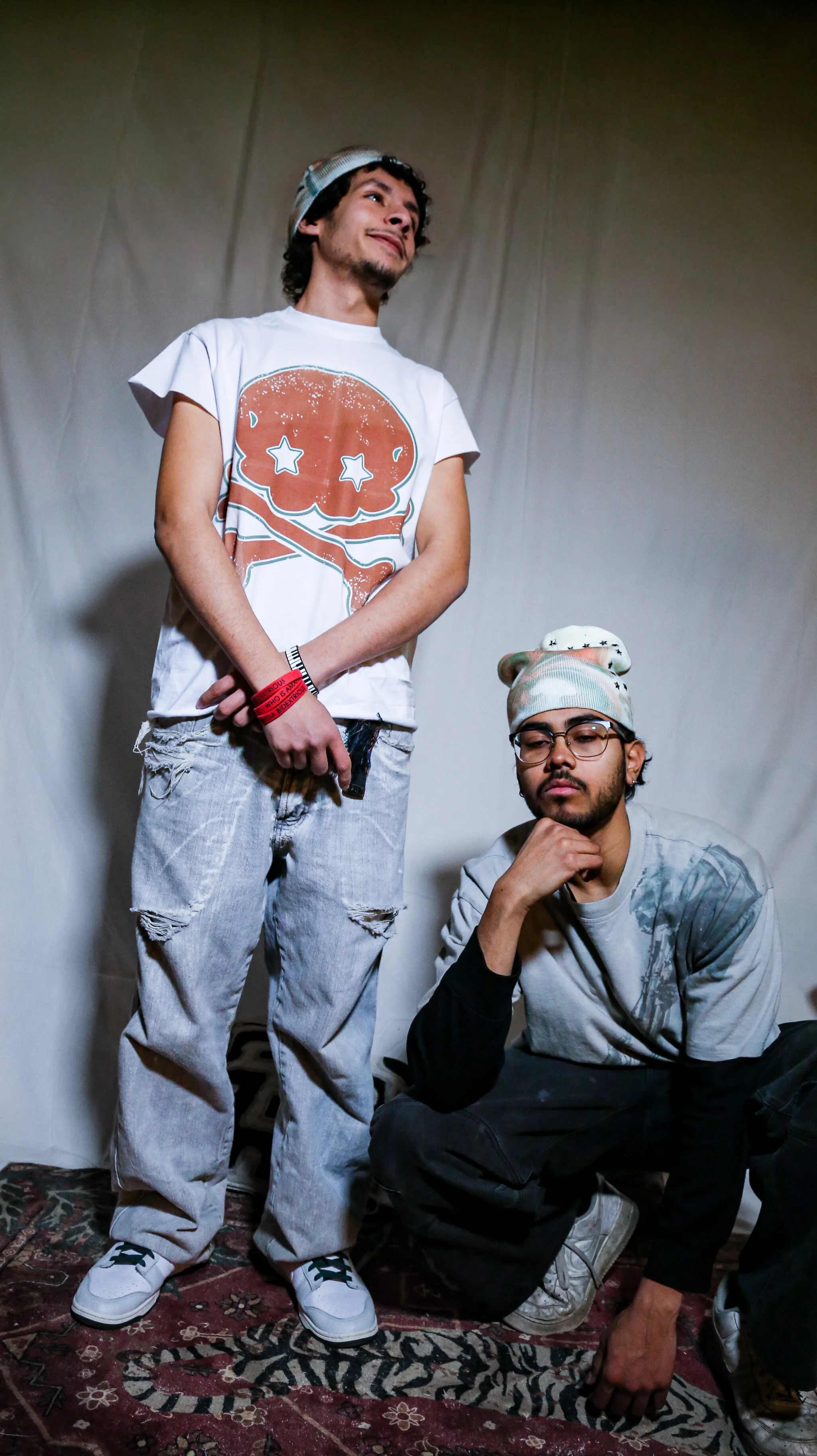 Two young men with casual clothing and accessories pose in front of a plain backdrop. One is standing, and the other is crouching.