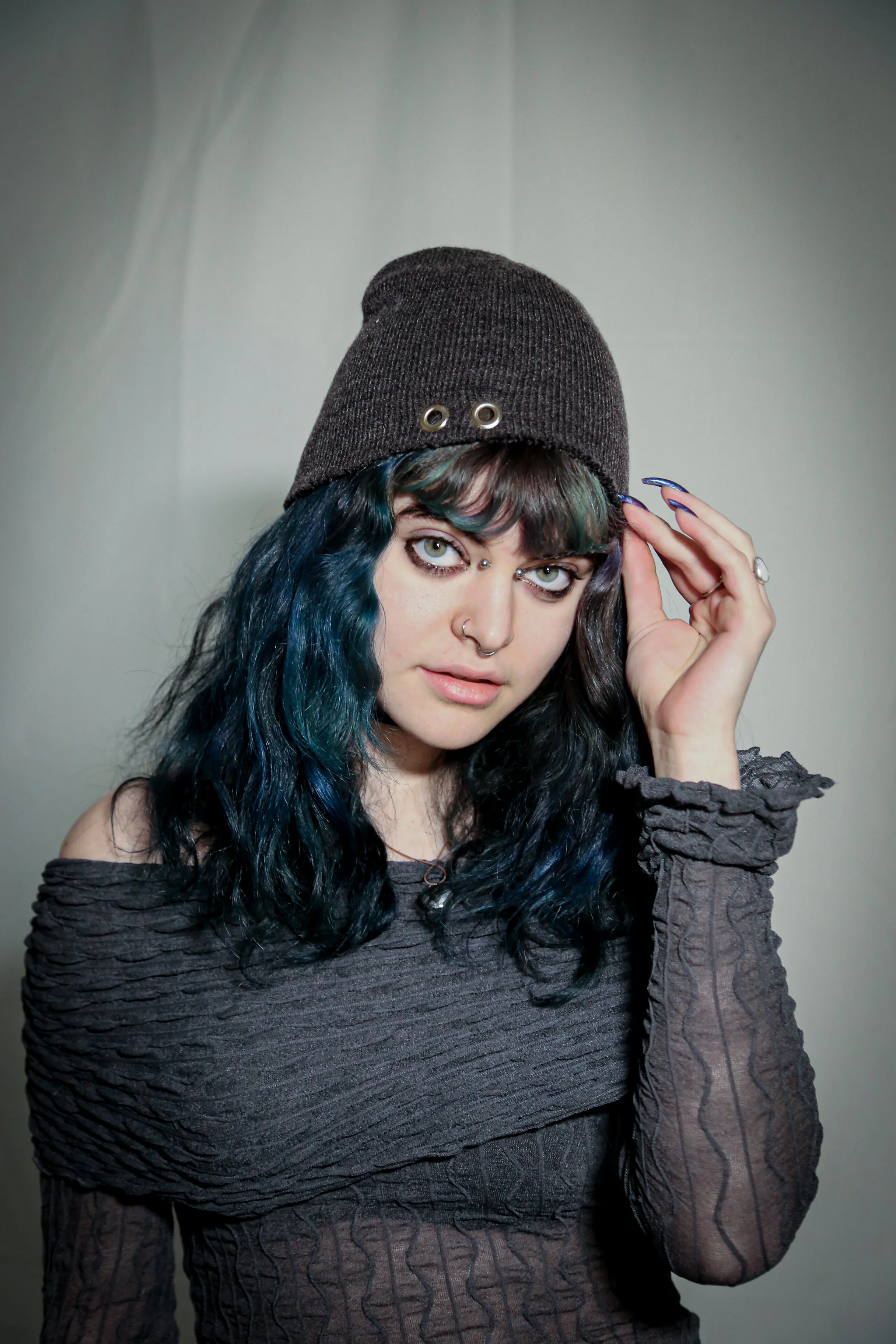 A young woman with curly blue and black hair wearing a dark gray beanie, black off-shoulder top, and silver jewelry, looking at the camera and touching her beanie.