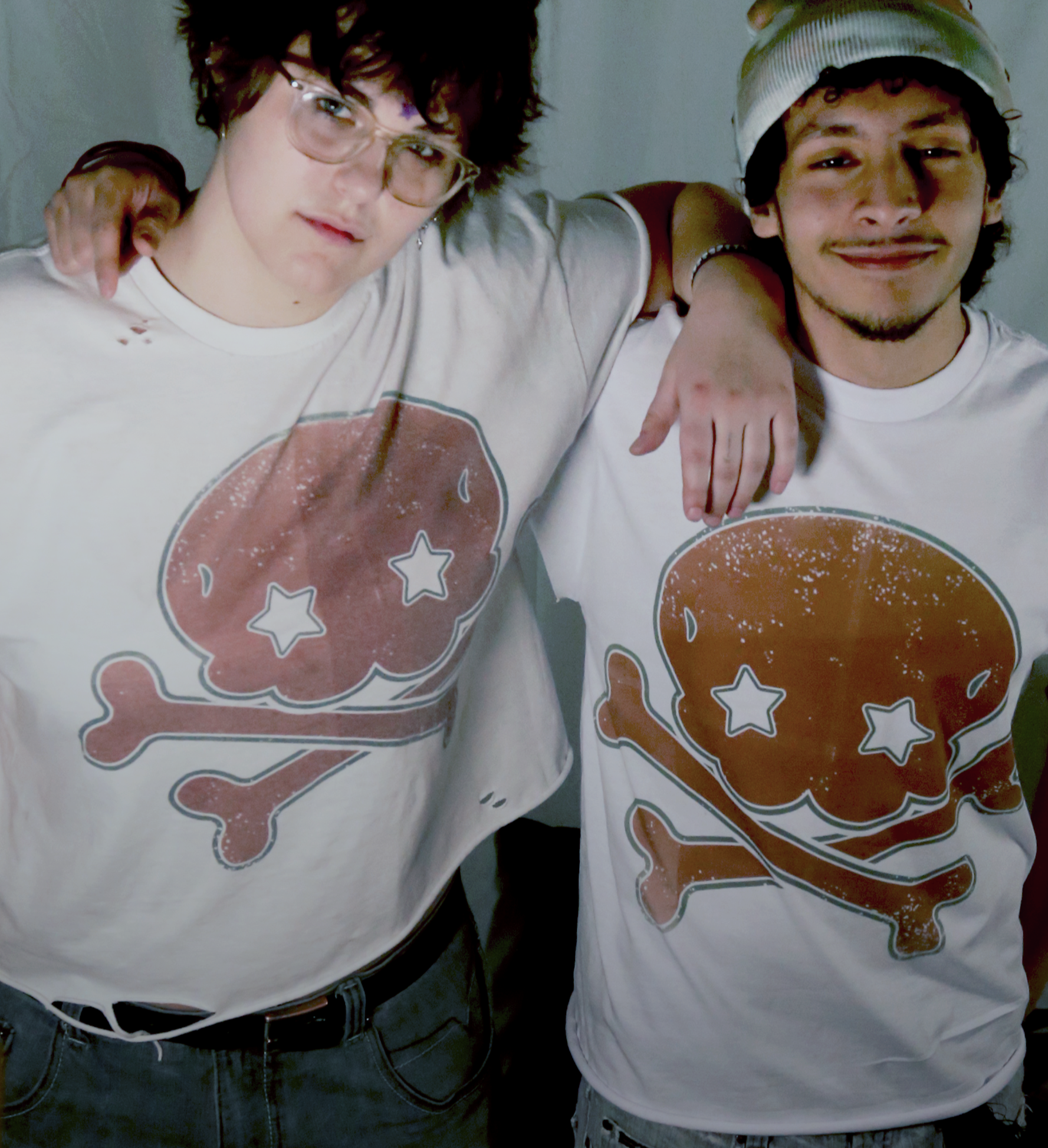 Two young men standing close together with their arms around each other's shoulders, wearing matching white T-shirts with a graphic of a skull and crossbones featuring star-shaped eyes.