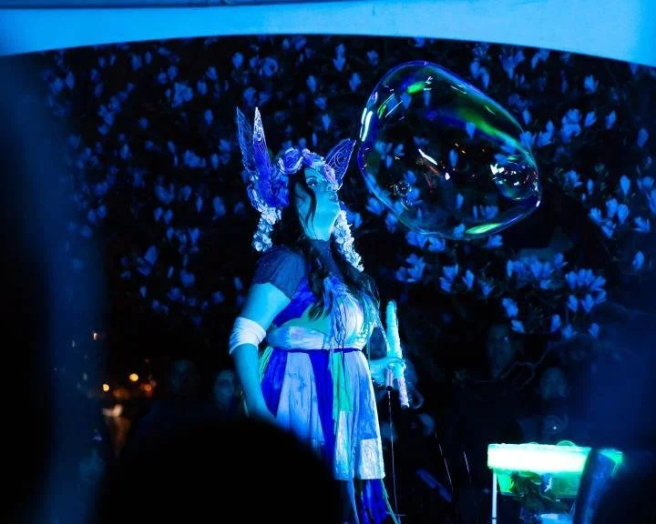 A performer dressed as a fantasy character with blue and purple costume, long dark hair, and a headpiece with ears and antlers, blowing a large iridescent bubble in front of an audience.