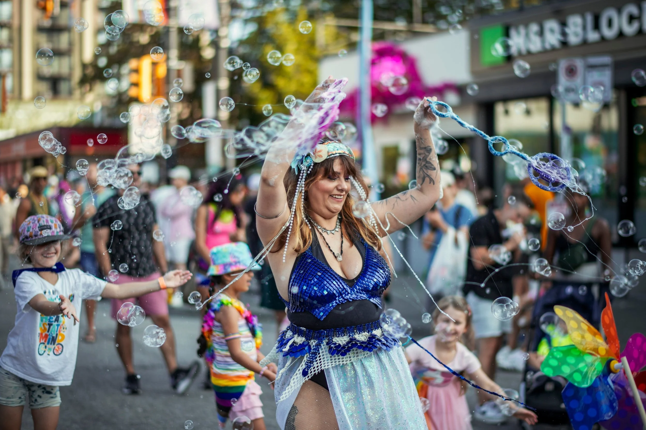 A woman with tattoos in a blue costume and a headpiece is blowing bubbles while children dance around her at a lively street festival, with colorful decorations and onlookers in the background.