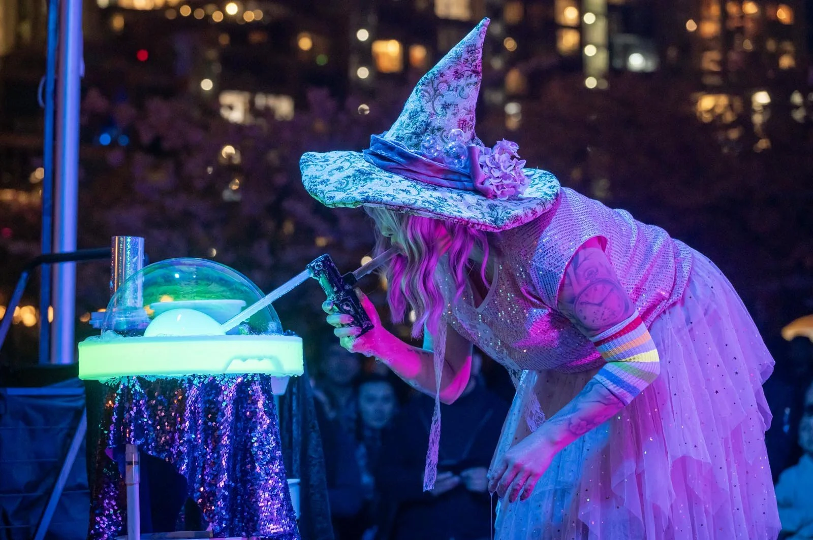 A person dressed as a witch in a pink sparkly dress and a tall, decorated hat, sipping a drink from a straw at an event with colorful lighting.