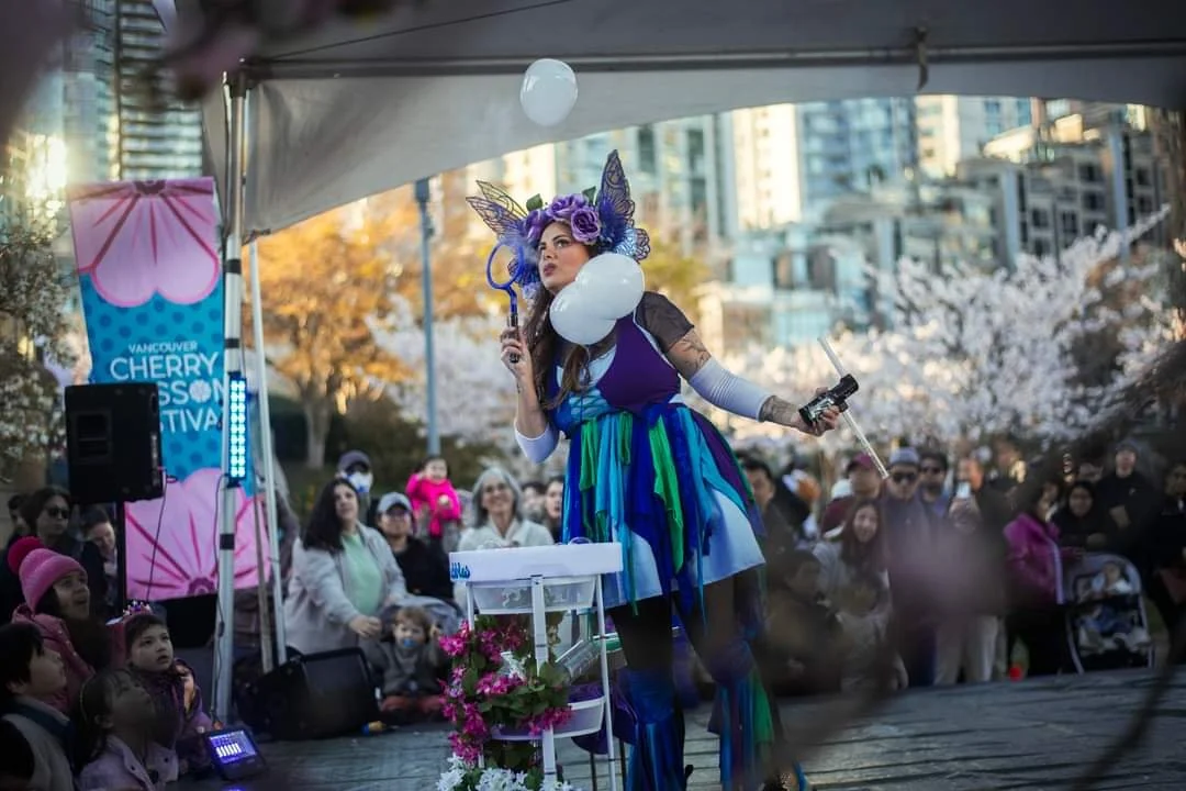 Performer dressed as a fairy with butterfly wings, a purple and blue dress, and a floral headpiece, performing on an outdoor stage at Vancouver Cherry Blossom Festival, surrounded by an audience in an urban park with cherry blossoms.
