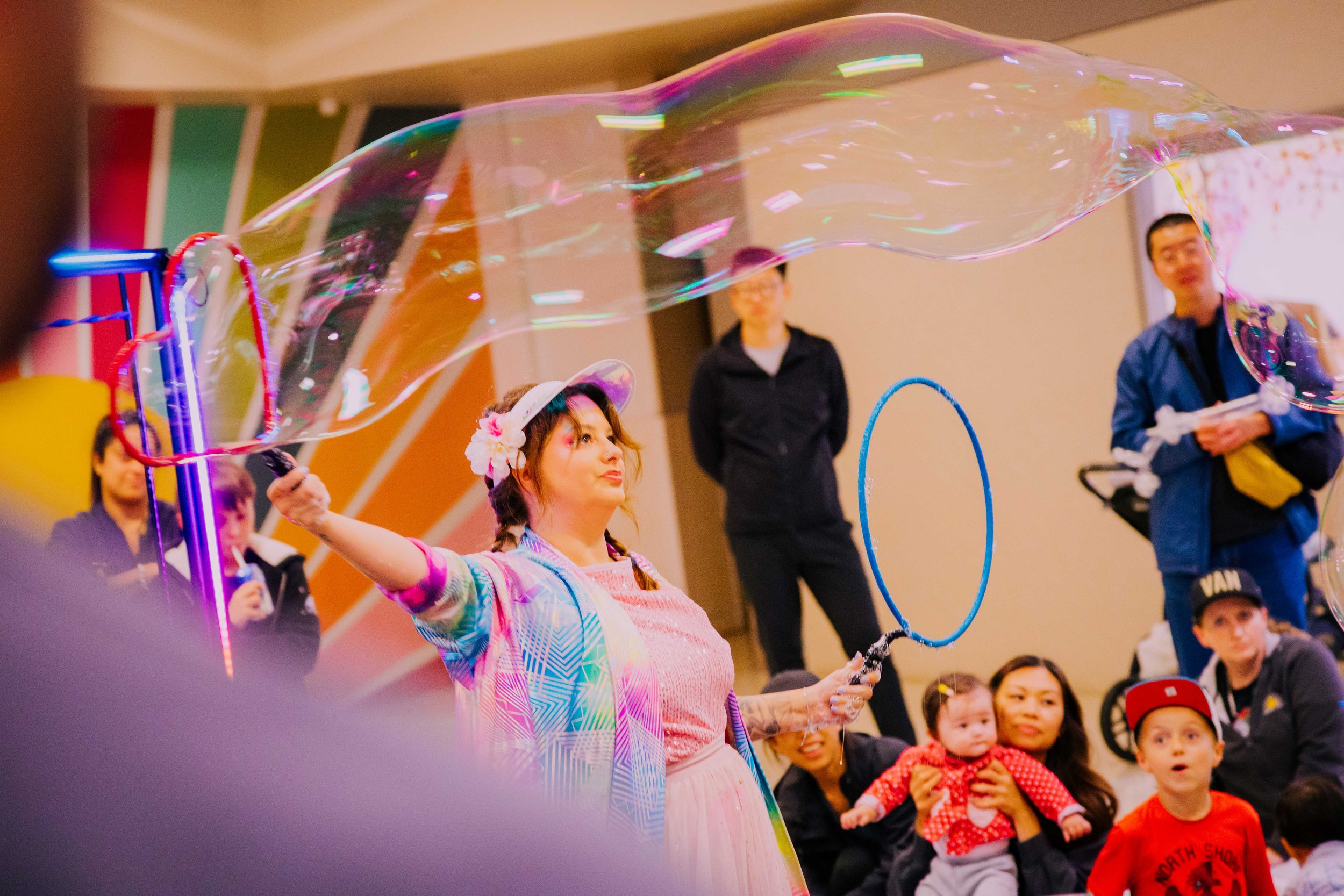 A woman with a hat and colorful outfit creates a large soap bubble with a hoop, surrounded by children and adults at an indoor event.