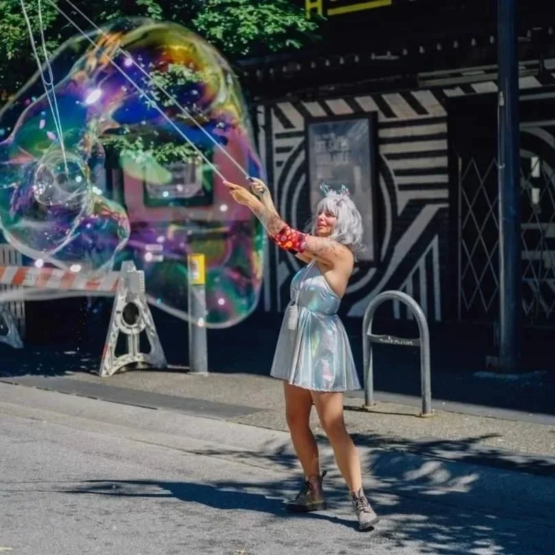 A woman with short, silver hair wearing a shiny, silver dress creating a large soap bubble on a city street.