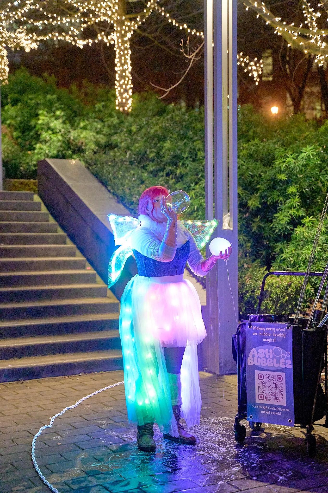 Person in a colorful, illuminated costume with glowing fairy wings, drinking from a glass, standing next to a cart with a QR code. The scene is outdoors at night with stairway and trees decorated with string lights in the background.