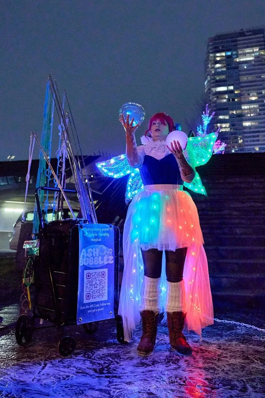 A performer dressed as a fairy with illuminated wings, holding two glowing orbs, standing outdoors at night in front of tall buildings, with a cart displaying a sign and QR code.