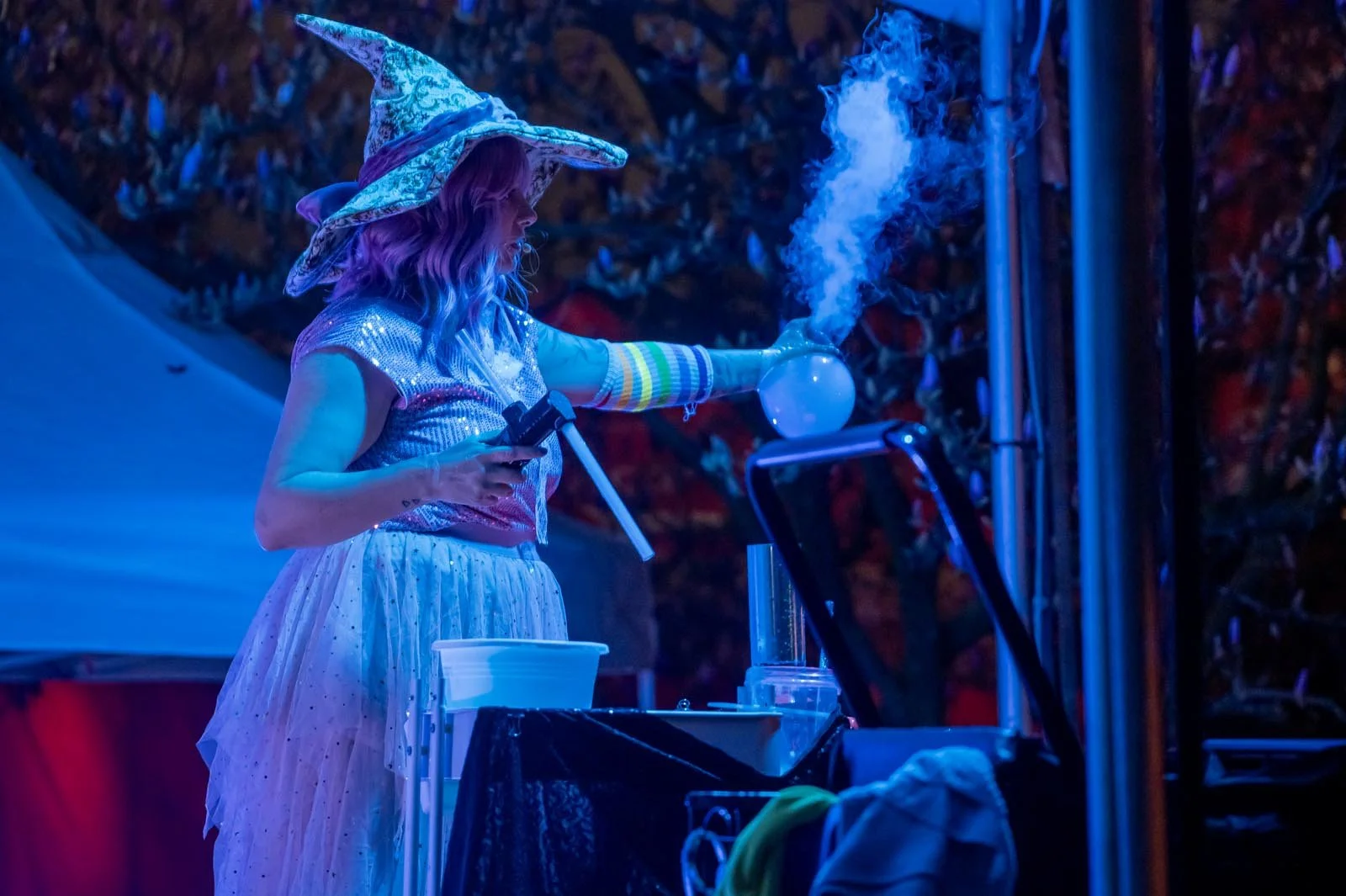 A woman dressed as a witch with a star-patterned hat and colorful arm warmers is blowing smoke from a round glass apparatus during a nighttime outdoor event, illuminated by blue lights.